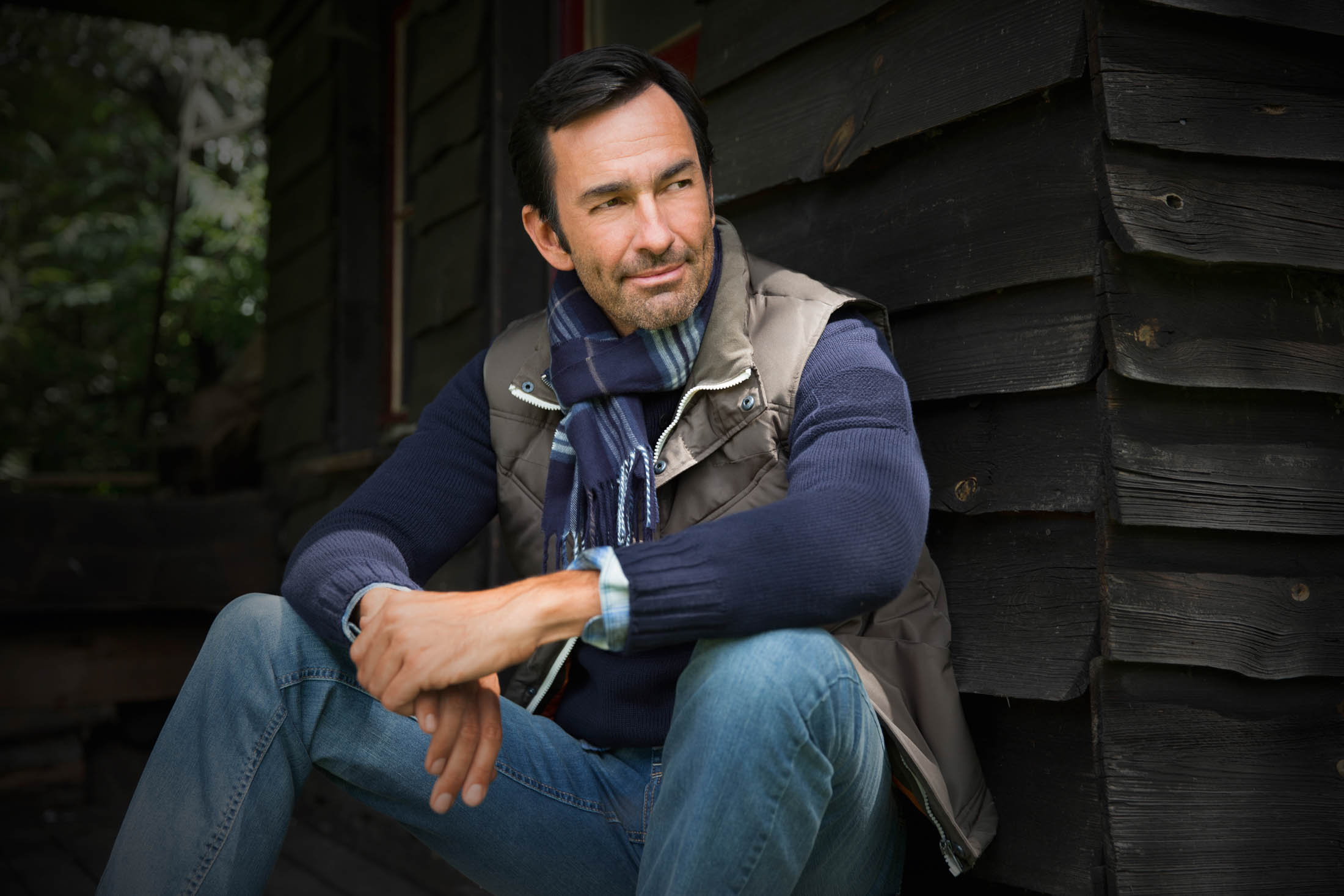 Ein Mann sitzt neben einer Holzhütte. Er trägt eine Weste, einen Schal und Jeans. Er sieht nachdenklich aus, während er in die Ferne blickt und die Arme um ein angehobenes Knie geschlungen hat. © Fotografie Tomas Rodriguez