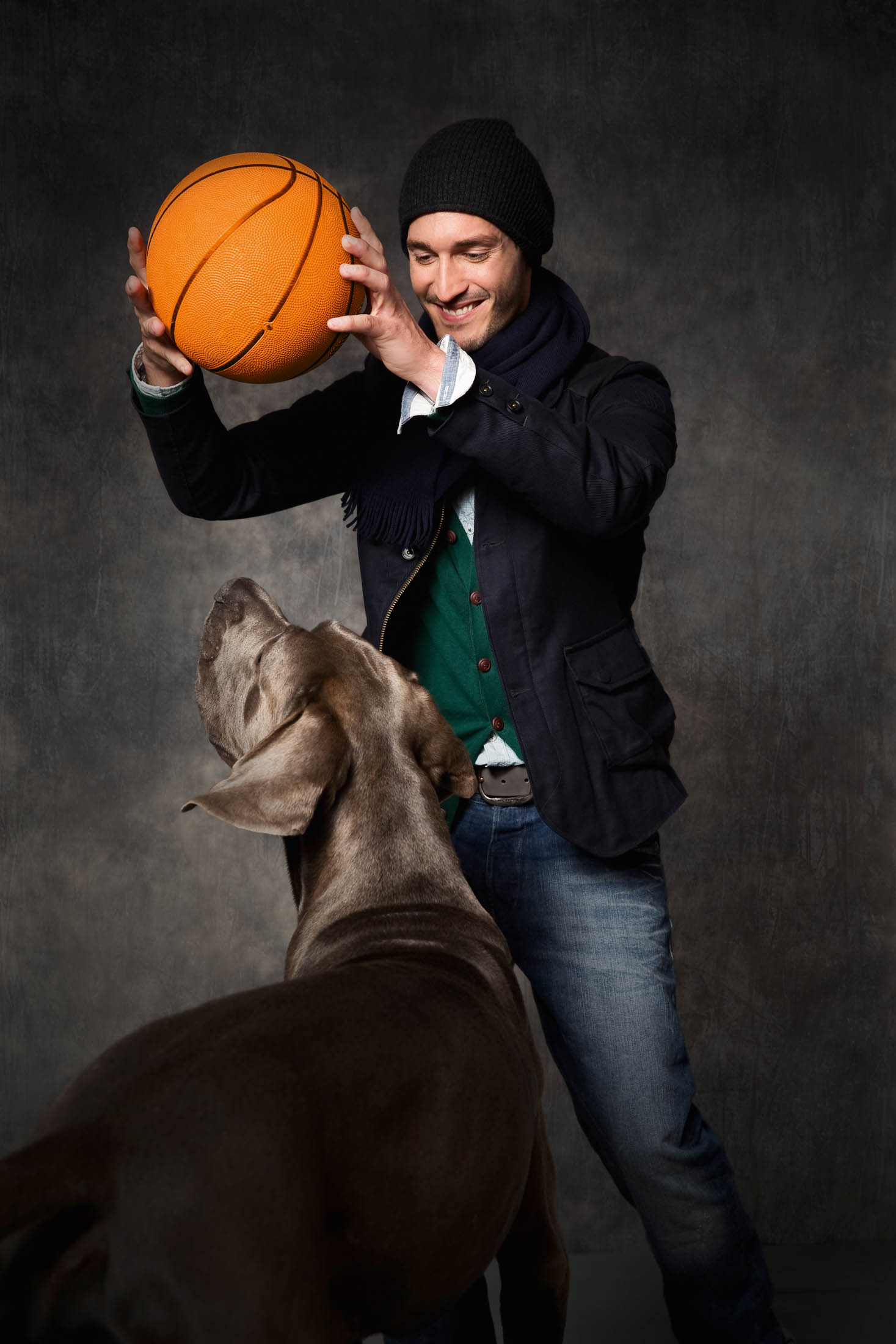 Vor einem dunkelgrauen Hintergrund hält ein Mann mit Mütze und Mantel einen Basketball über seinem Kopf und spielt spielerisch mit einem großen grauen Hund, der hochspringt, um ihn zu erreichen. © Fotografie Tomas Rodriguez