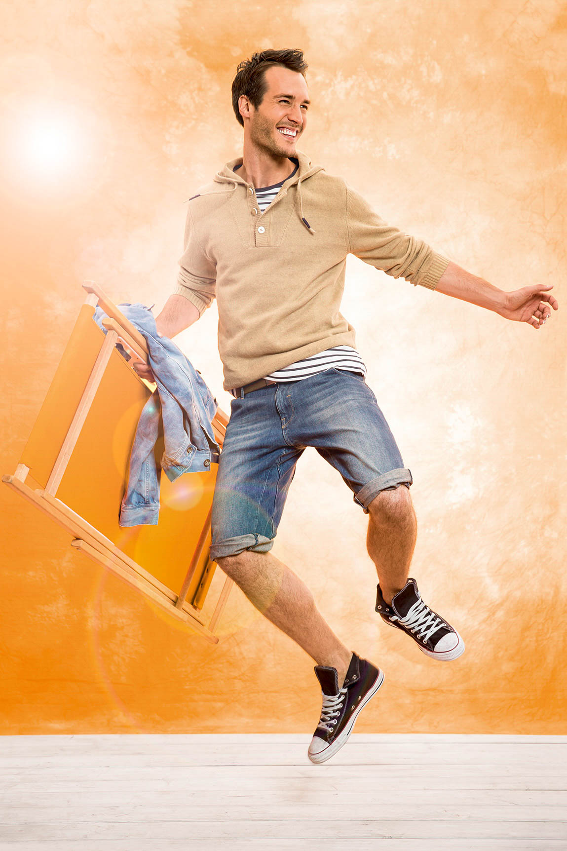 Ein fröhlicher Mann in einem beigen Kapuzenpullover und Jeansshorts springt neben einem orangefarbenen Stuhl herum, über den eine Jeansjacke hängt, vor einem warmen, strukturierten orangefarbenen Hintergrund. © Fotografie Tomas Rodriguez