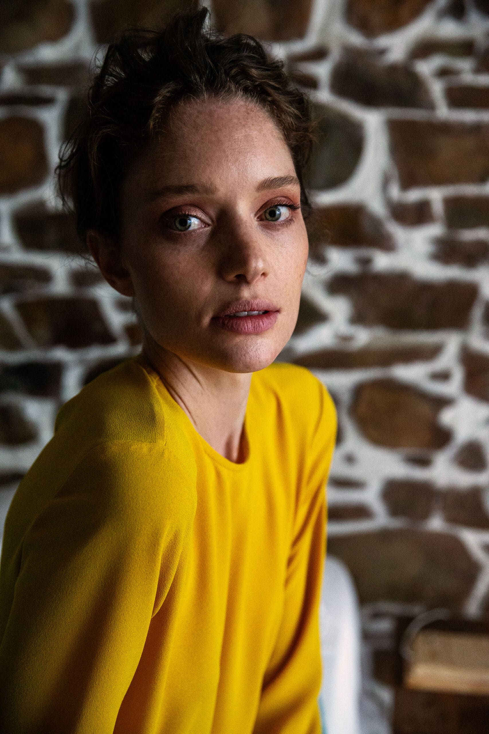 Eine Frau mit zerzaustem Haar und einer gelben Bluse sitzt vor einer rustikalen Backsteinmauer und blickt aufmerksam in die Kamera. © Fotografie Tomas Rodriguez