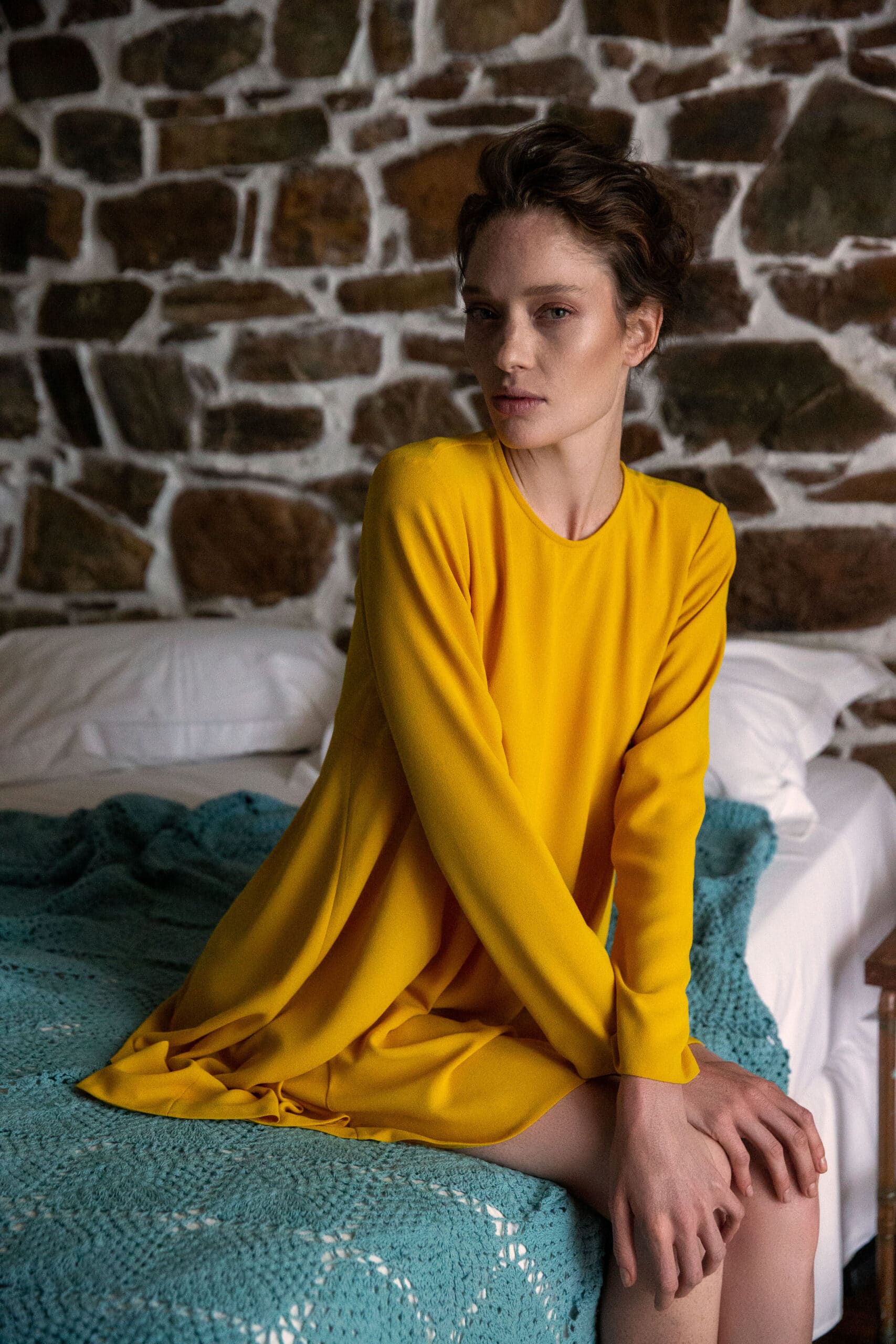 Eine Frau in einem gelben Kleid sitzt nachdenklich auf einer blauen Decke, im Hintergrund ist eine rustikale Steinmauer zu sehen. © Fotografie Tomas Rodriguez