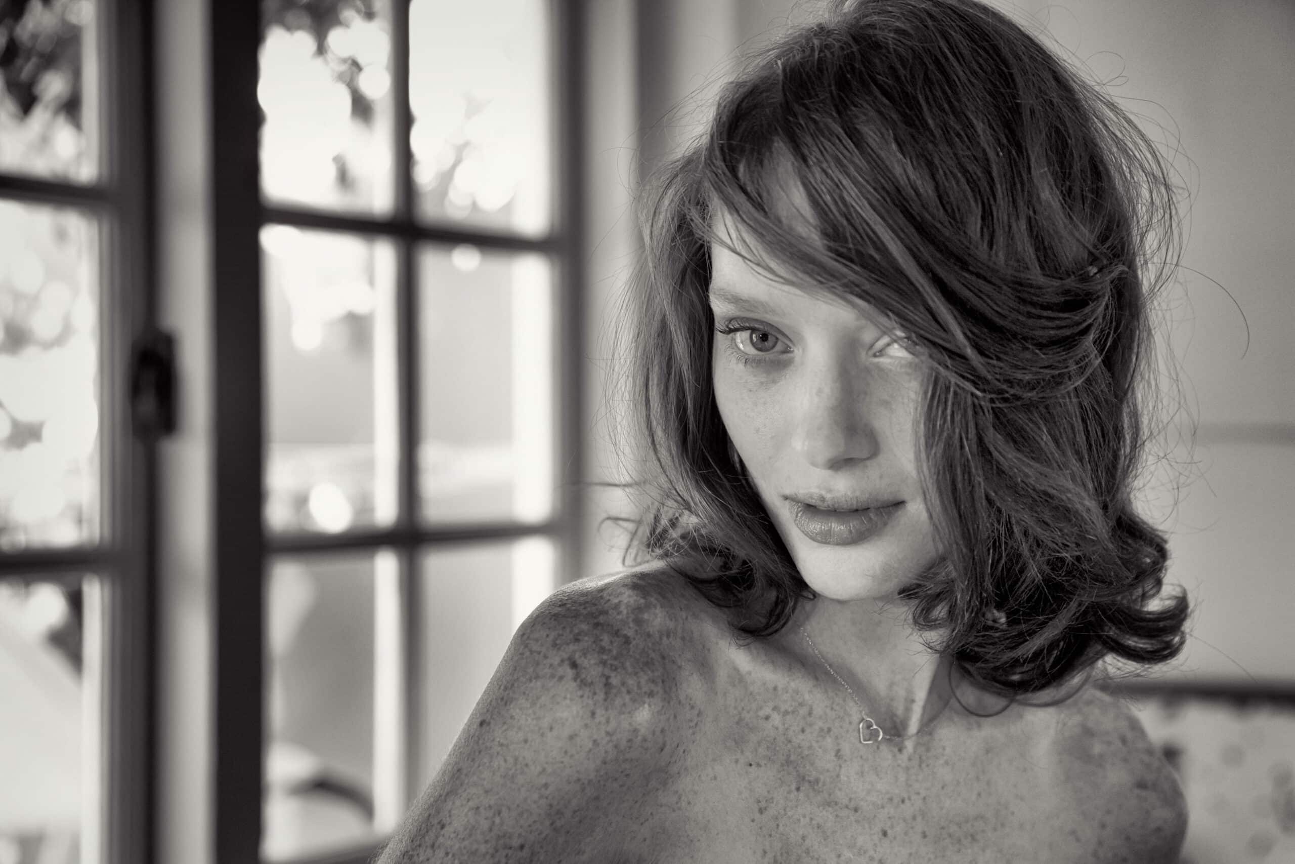 Schwarz-Weiß-Porträt einer Frau mit zerzaustem Haar, die mit aufmerksamem Blick in die Kamera blickt, neben einem gut beleuchteten Fenster mit Türen im Hintergrund. © Fotografie Tomas Rodriguez