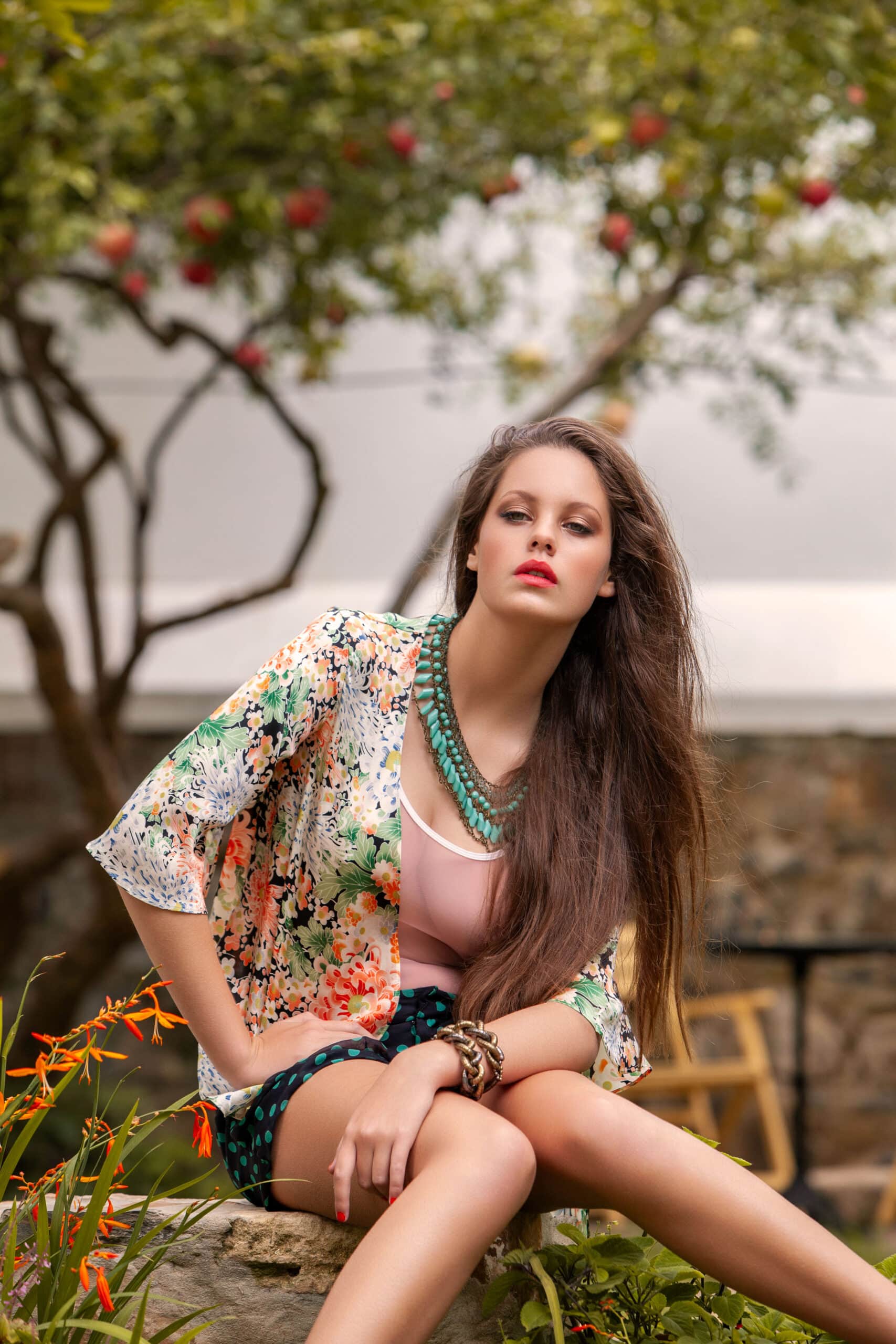 Eine Frau in einer geblümten Jacke und einem rosa Oberteil sitzt elegant auf dem Boden in einem Garten mit blühenden Blumen und üppigem Grün und blickt nachdenklich zur Seite. © Fotografie Tomas Rodriguez