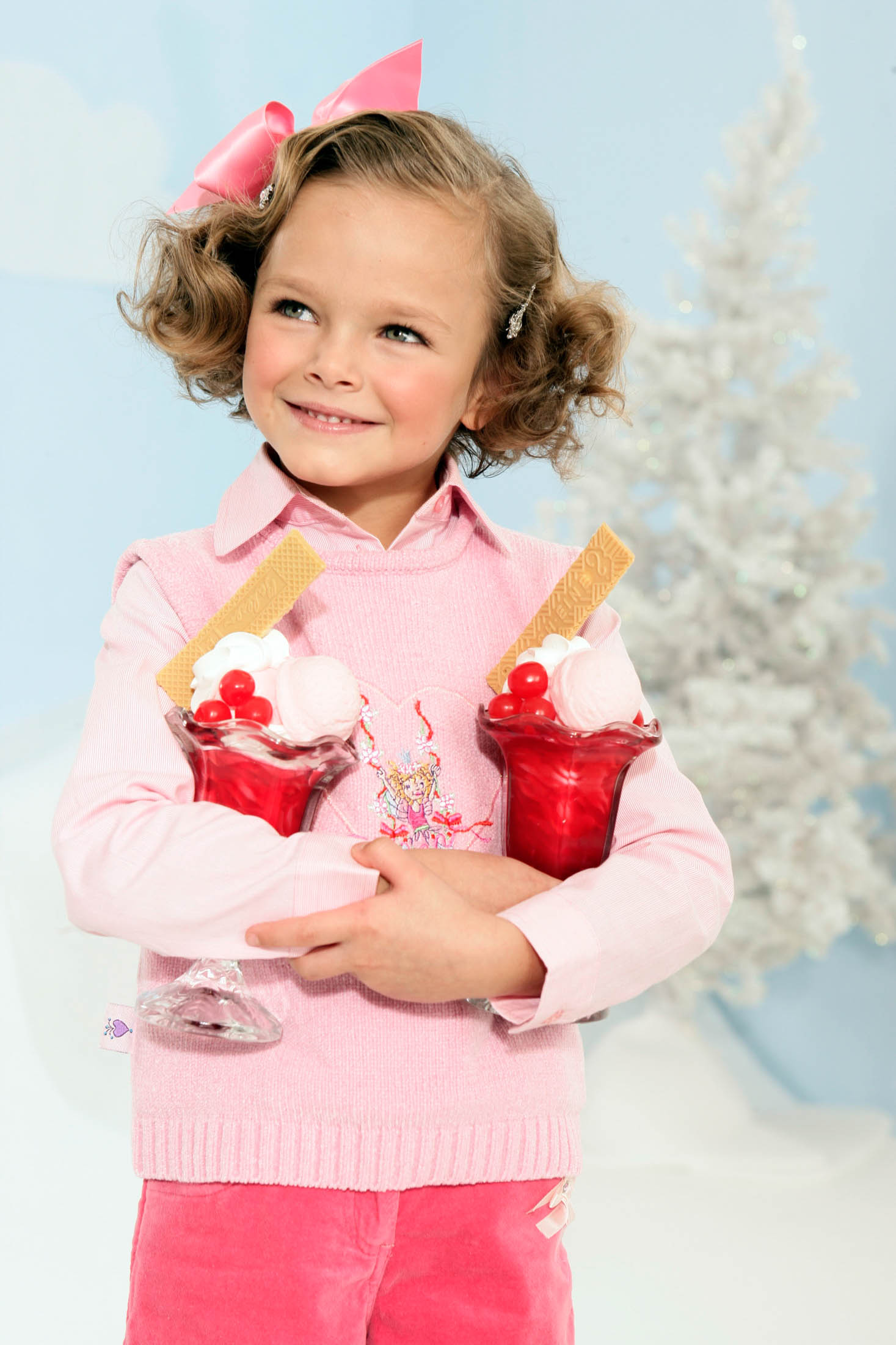 Ein fröhliches junges Mädchen mit lockigem Haar, einem rosa Pullover und zwei eistütenförmigen Leckereien in der Hand lächelt breit vor dem Hintergrund eines hellblauen Himmels und eines verschwommenen Weihnachtsbaums. © Fotografie Tomas Rodriguez