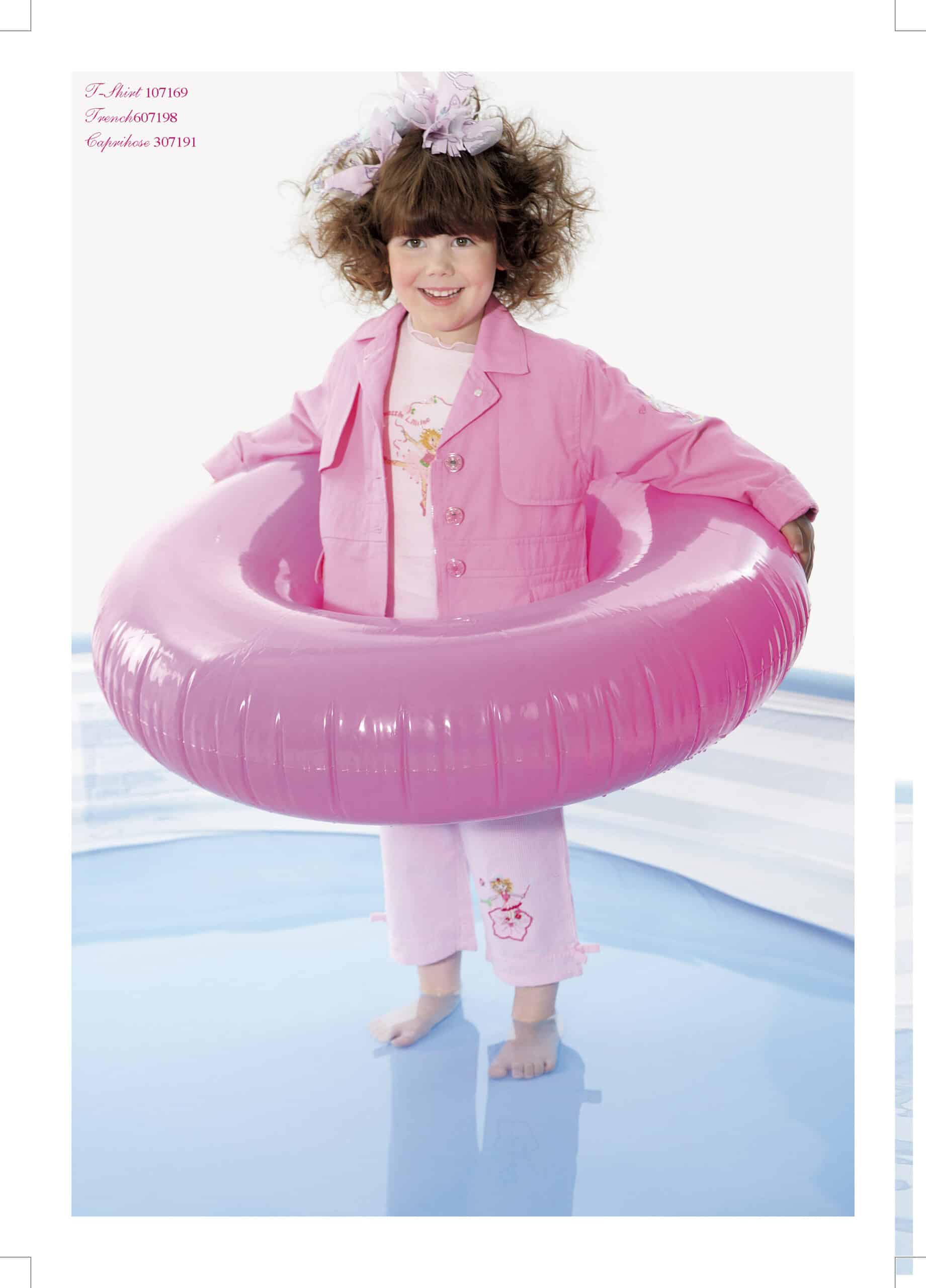 Ein junges Mädchen in rosa Jacke und Hose steht lächelnd in einem flachen Pool, trägt einen rosa aufblasbaren Ring um die Hüfte und hat lockiges braunes Haar. Als Accessoire trägt sie eine große rosa Blume. © Fotografie Tomas Rodriguez