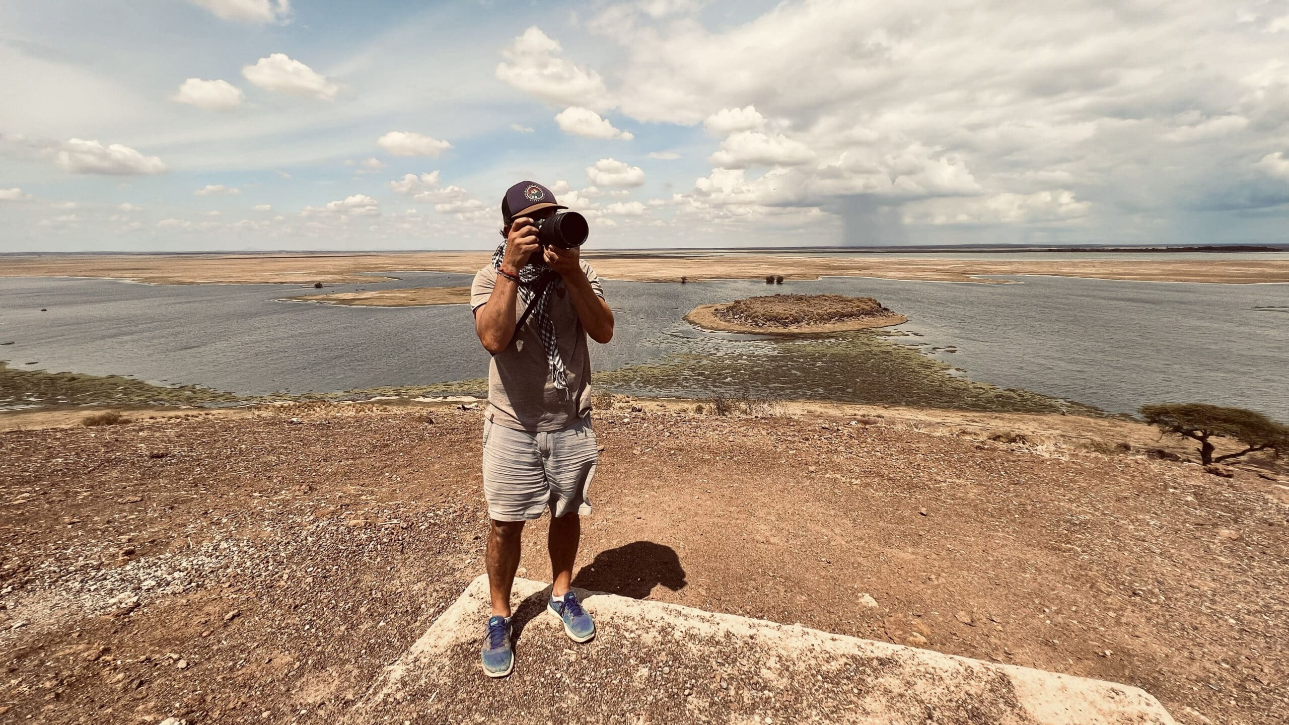 Ein Fotograf steht auf einem felsigen Aussichtspunkt und macht mit einer DSLR-Kamera ein Foto. Im Hintergrund sind ausgedehnte Feuchtgebiete und spärliche Bäume unter einem weiten, wolkenverhangenen Himmel zu sehen. © Fotografie Tomas Rodriguez