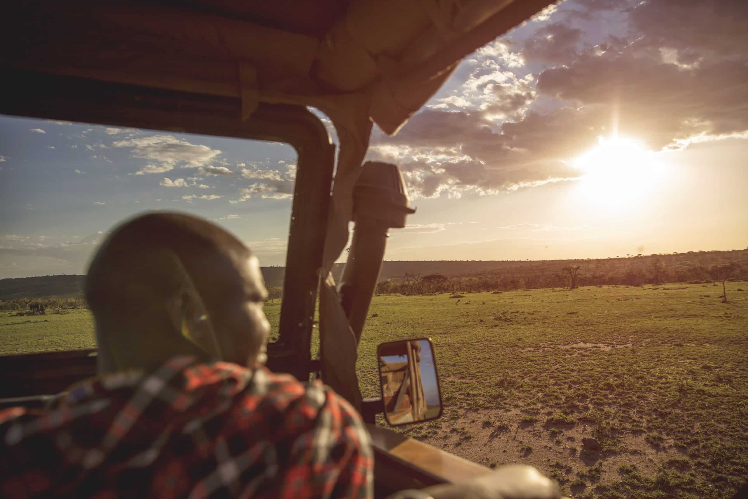 Eine Person im karierten Hemd beobachtet von einem Safarifahrzeug aus den Sonnenuntergang und hat Blick auf eine weite Savannenlandschaft. © Fotografie Tomas Rodriguez