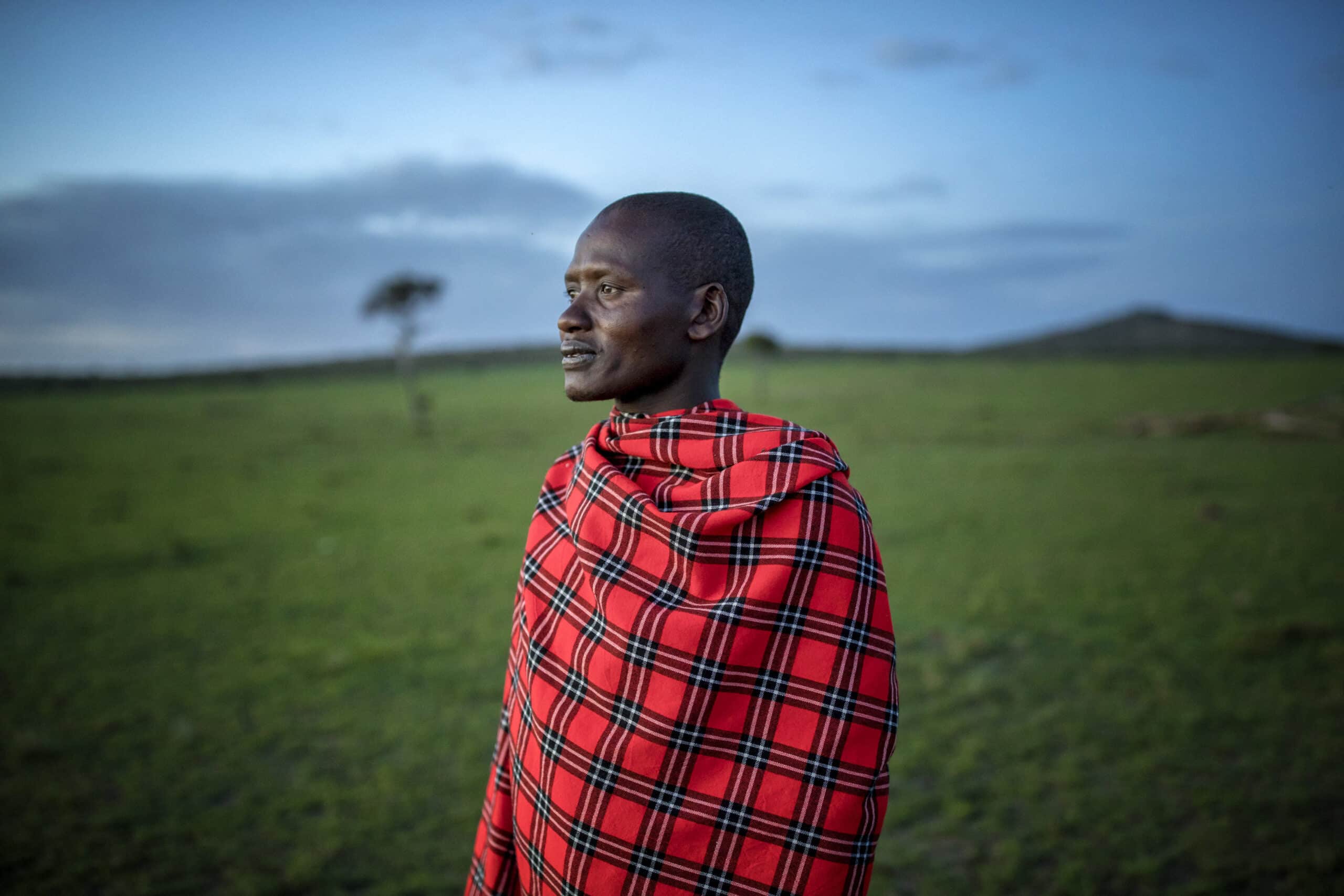 Ein Mann in traditioneller Massai-Kleidung, mit einem rot-blauen Shuka, steht in der Abenddämmerung auf einer Wiese und blickt mit heiterem Gesichtsausdruck zur Seite. Im Hintergrund sind sanft verschwommene Bäume und ein Hügel zu sehen. © Fotografie Tomas Rodriguez