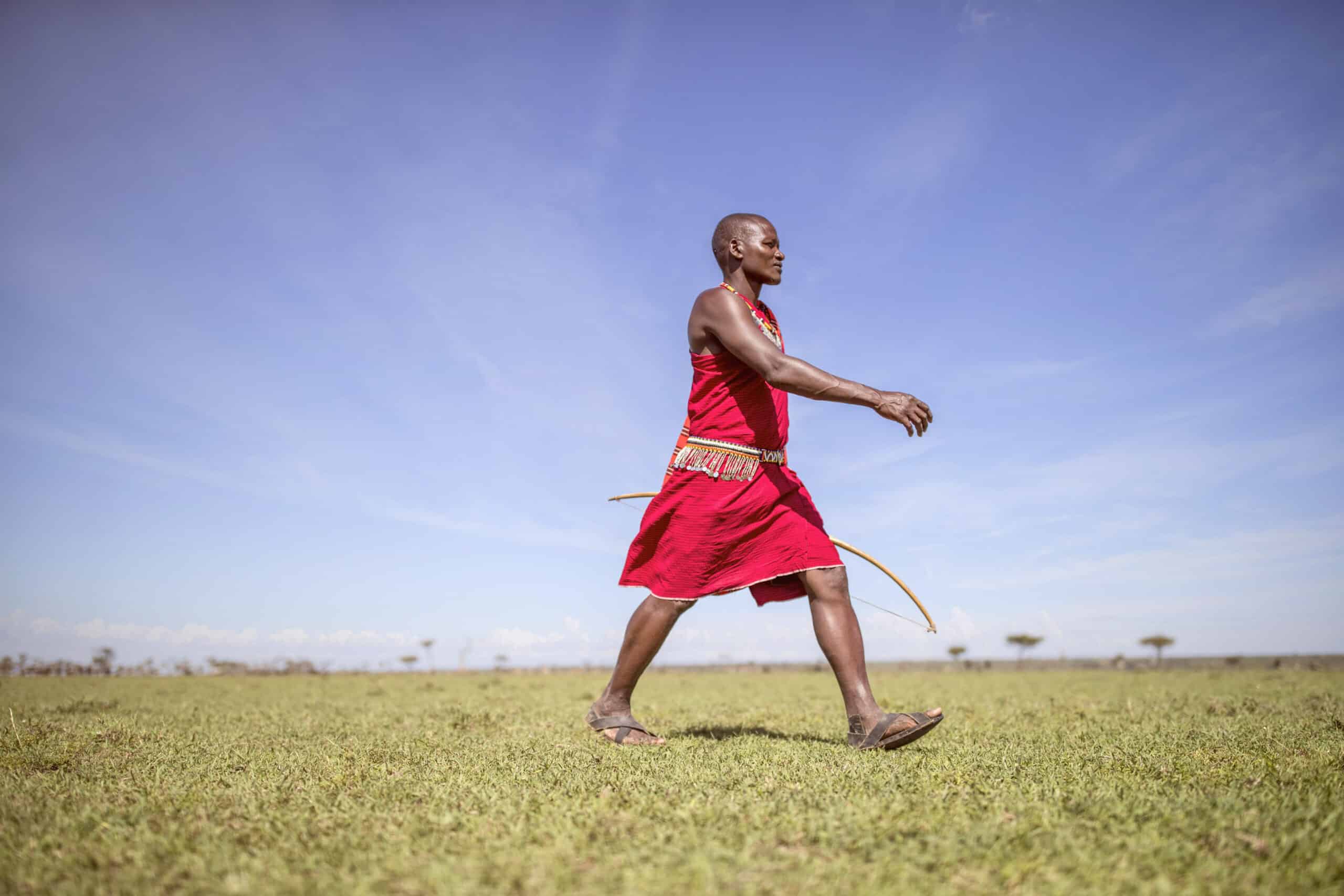 Ein Massai-Mann in traditioneller Kleidung geht unter einem klaren blauen Himmel über eine Wiese und hält in entspannter Haltung einen Bogen. © Fotografie Tomas Rodriguez