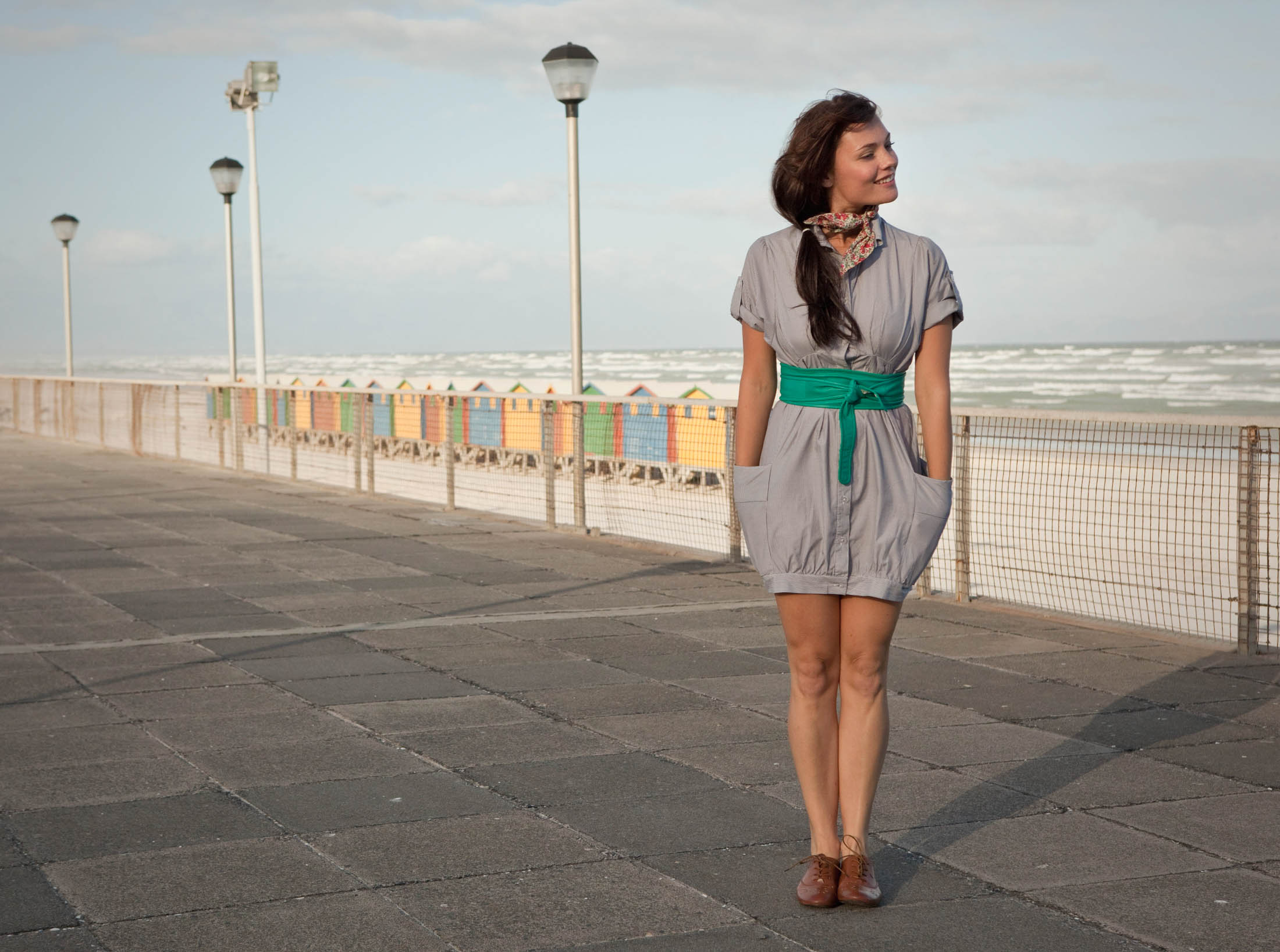 Eine Frau steht auf einer Strandpromenade, blickt mit einem leichten Lächeln nach links, sie trägt ein graues Kleid und einen grünen Gürtel. Im Hintergrund sind bunte Strandhütten und starke Meereswellen unter einem bewölkten Himmel zu sehen. © Fotografie Tomas Rodriguez