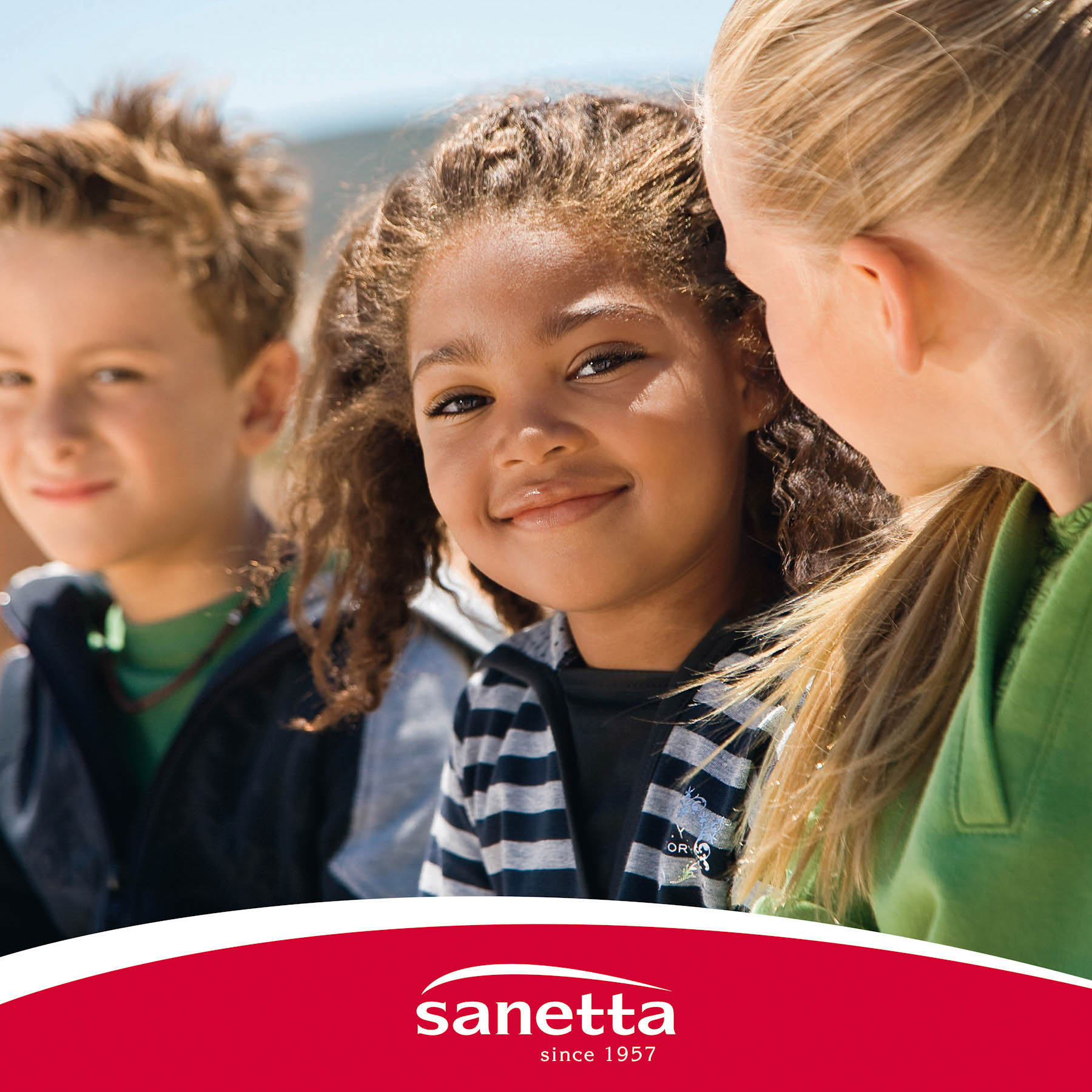 Drei Kinder lächeln im Freien; in der Mitte blickt ein Mädchen direkt in die Kamera, auf jeder Seite zwei Jungen, von denen einer teilweise sichtbar ist. Darunter ist das Logo von Sanetta zu sehen. Im Hintergrund ein heller, sonniger Tag. © Fotografie Tomas Rodriguez