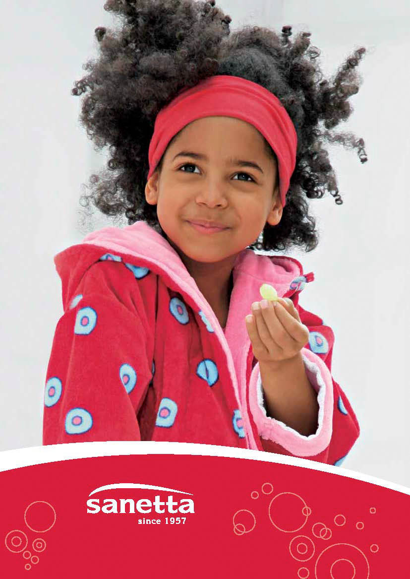 Ein fröhliches junges Mädchen mit lockigem Haar und einem roten Stirnband, das einen rosa Bademantel mit blauen Punkten trägt, lächelt und hält einen kleinen Gegenstand. Der Hintergrund ist schlicht weiß. © Fotografie Tomas Rodriguez