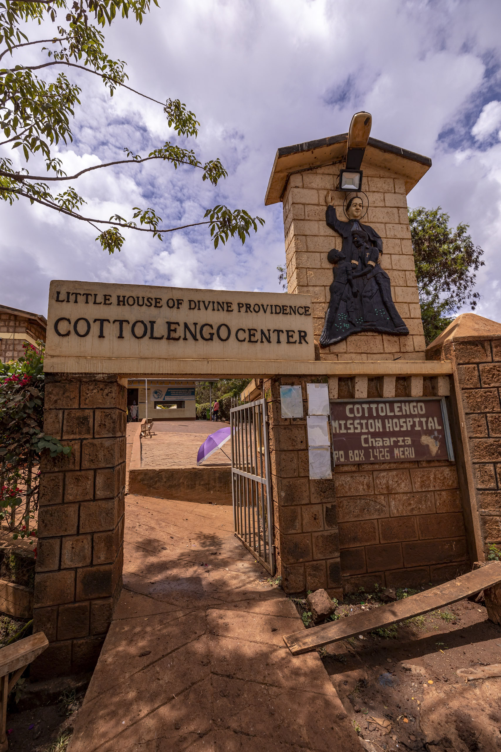 Ein Steintor mit einem Schild mit der Aufschrift „Little House of Divine Providence Cottolengo Center“ und „Cottolengo Mission Hospital, Chaaria“ vor einem Gebäude. Der Eingang verfügt über eine Laterne an der Decke und eine Skulptur einer religiösen Figur, die auf der Säule montiert ist. © Fotografie Tomas Rodriguez