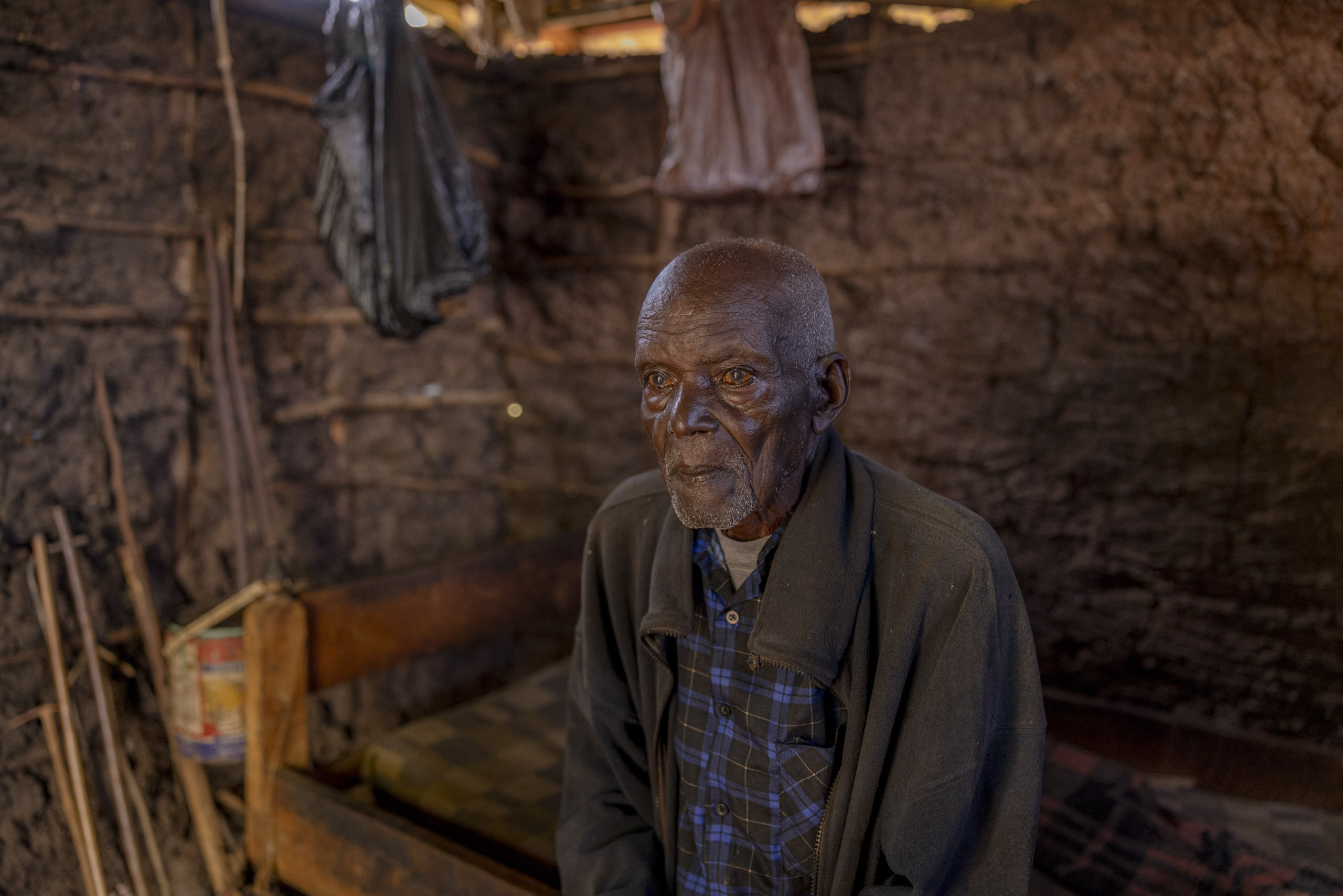 Ein älterer Mann mit nachdenklichem Gesichtsausdruck sitzt auf einer Holzbank in einer rustikalen Hütte mit Wänden aus Stöcken und Lehm. Seine Umgebung ist nur minimal möbliert und die Beleuchtung ist schwach. © Fotografie Tomas Rodriguez