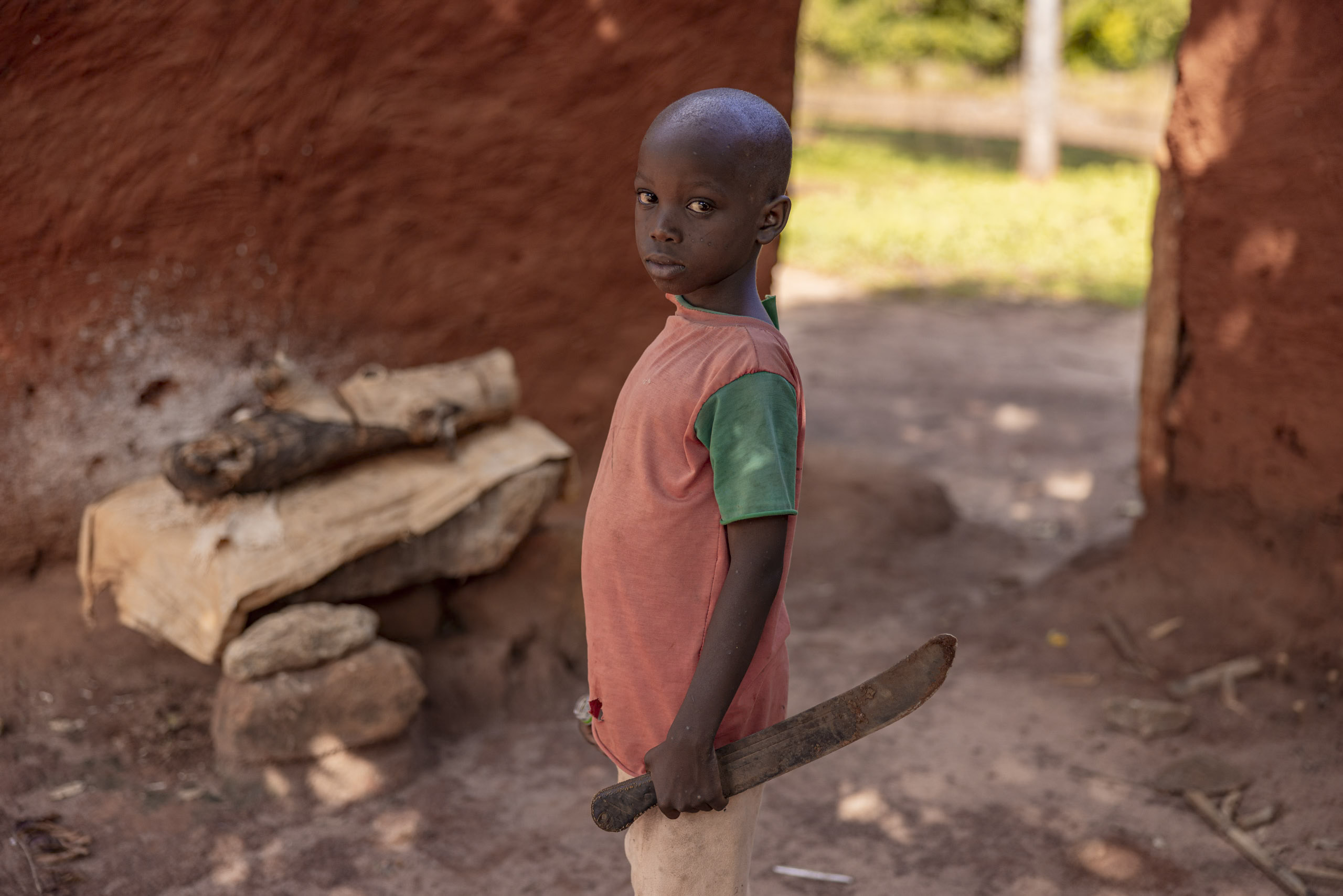 Ein kleiner Junge in einem rot-grünen Hemd hält eine Machete und steht in einem staubigen Dorf mit roten Lehmwänden und einer Holzbank im Hintergrund. © Fotografie Tomas Rodriguez