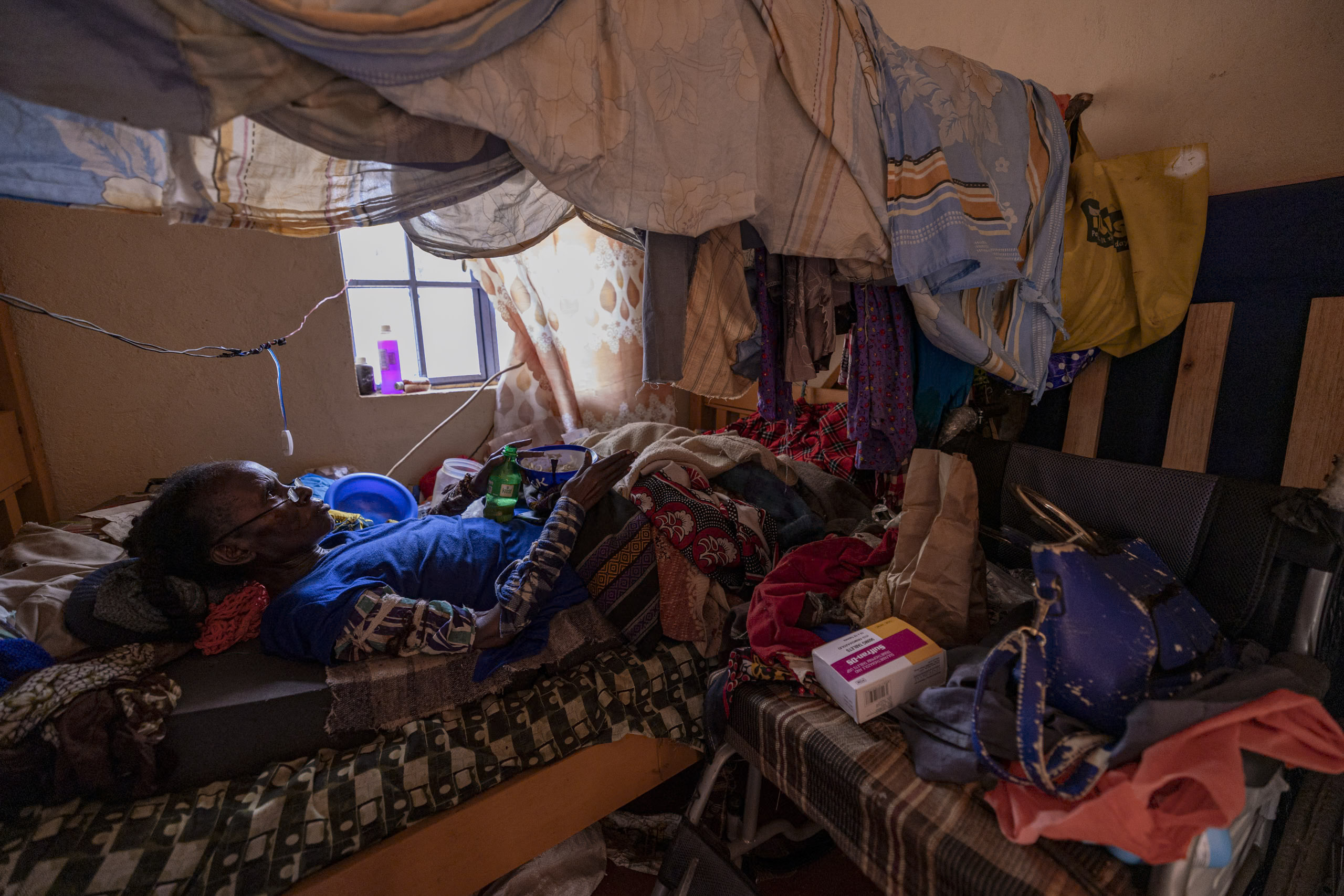 Ein Mann liegt in einem überfüllten Raum unter einem provisorischen Etagenbett, das mit verschiedenen Tüchern bedeckt ist, umgeben von persönlichen Gegenständen und vom Sonnenlicht, das durch ein Fenster einfällt. © Fotografie Tomas Rodriguez