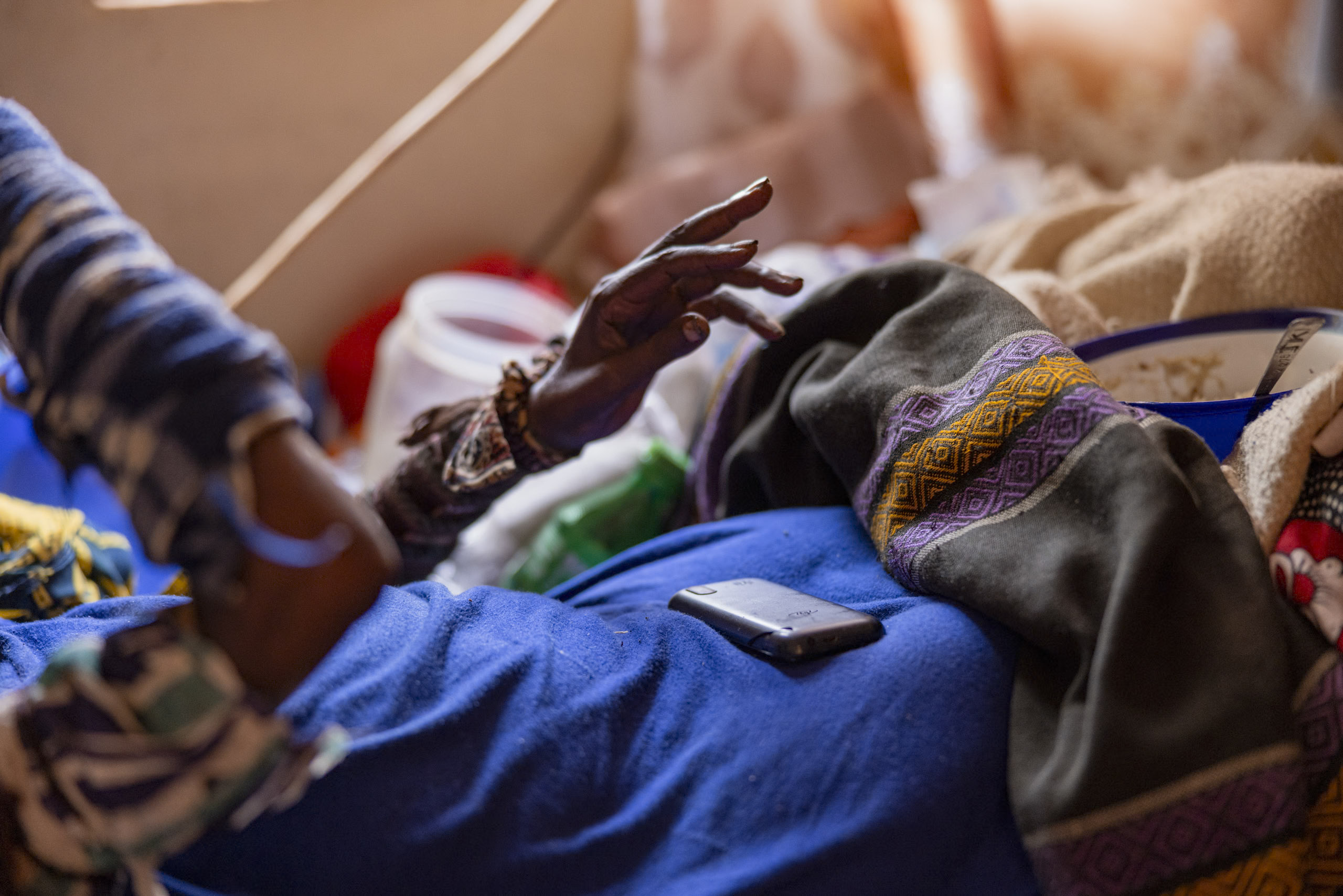 Eine Nahaufnahme der Hände einer Person, die an einem Textil arbeitet, mit buntem Stoff und einem Smartphone in der Nähe, in einem warm beleuchteten Raum. © Fotografie Tomas Rodriguez