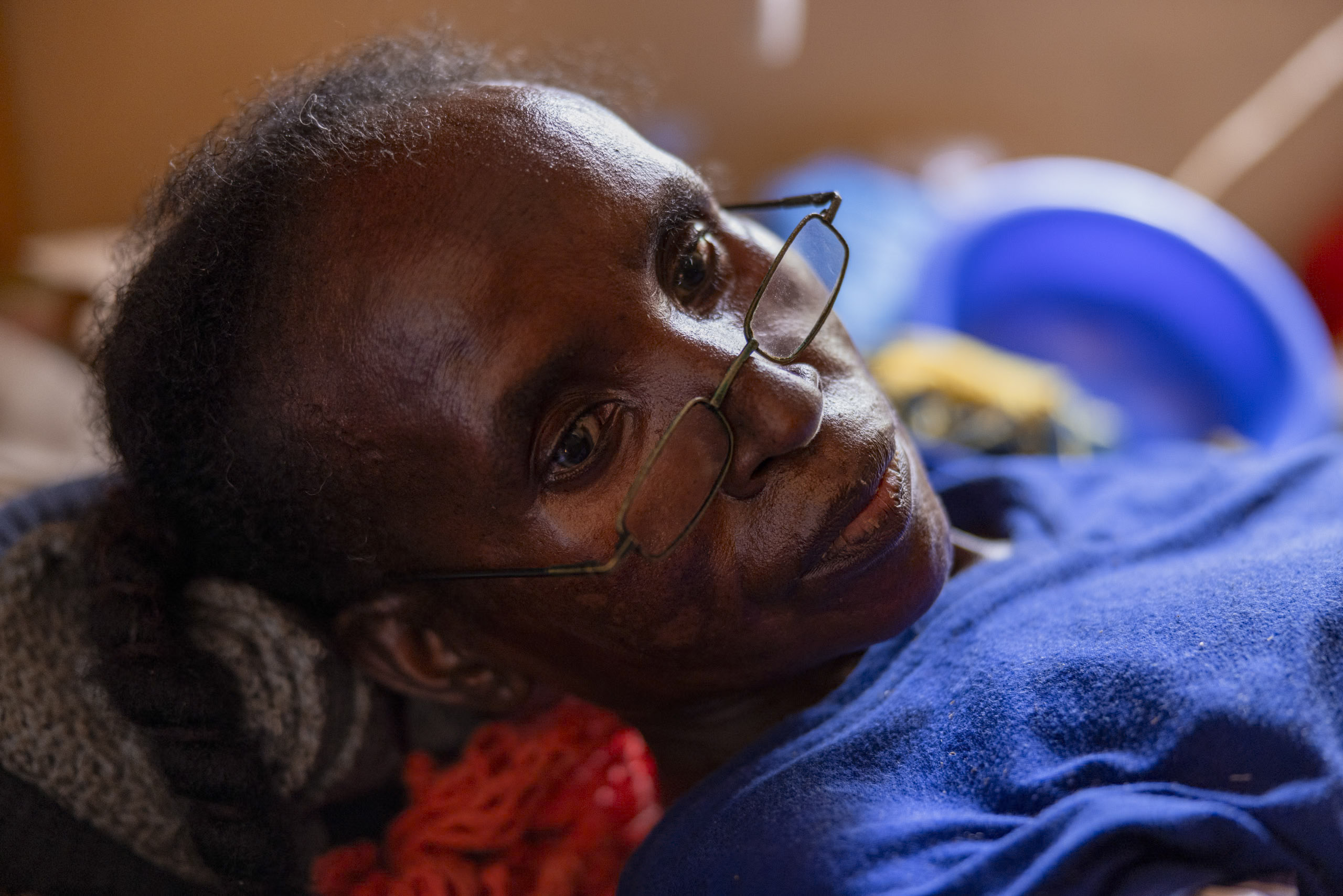 Eine ältere Frau mit Brille liegt auf der Seite und blickt nachdenklich in die Kamera, umgeben von einer warmen, intimen Innenatmosphäre. © Fotografie Tomas Rodriguez