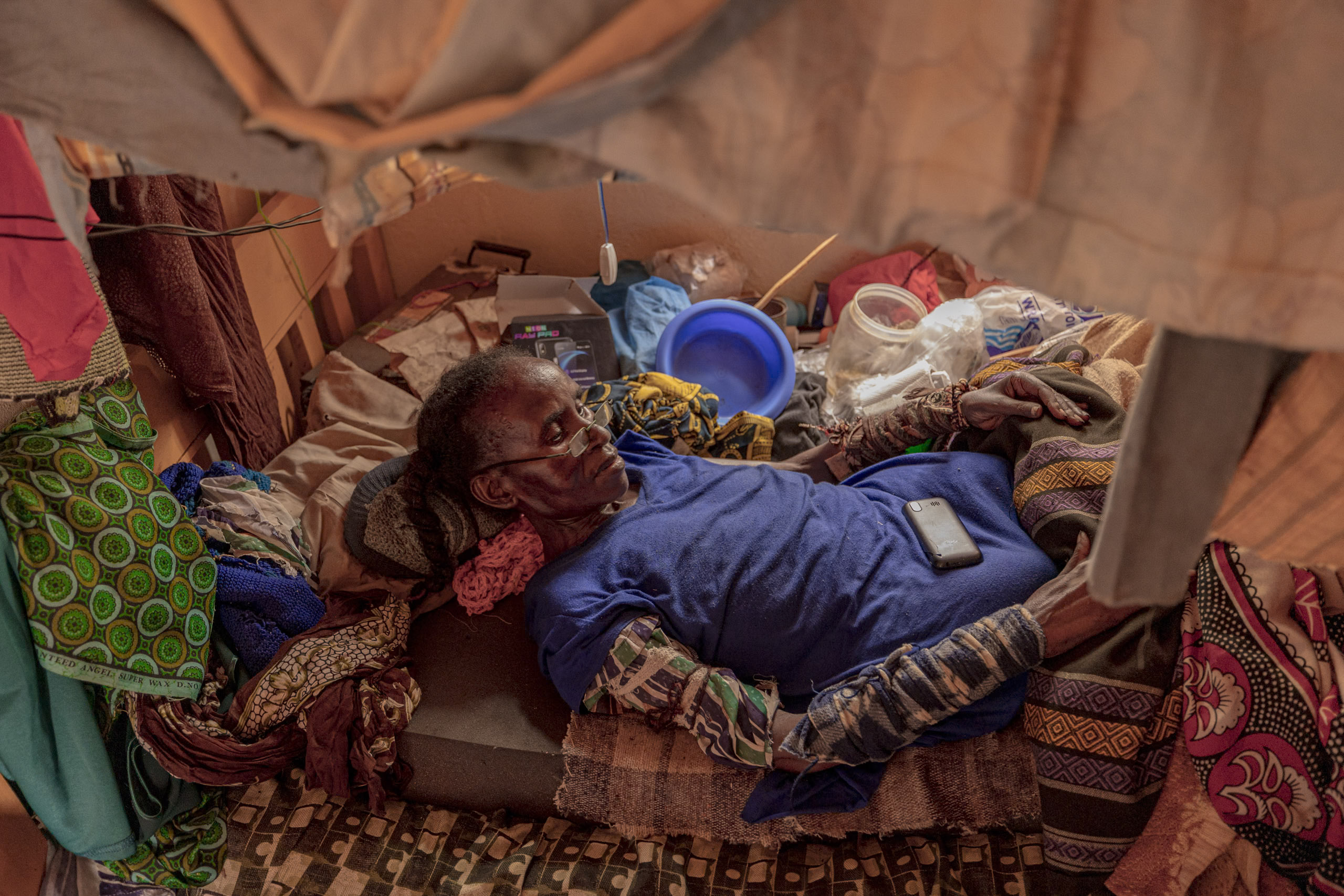 Eine ältere Frau liegt auf einem provisorischen Bett in einem engen Zelt, umgeben von persönlichen Gegenständen und einem Eimer. Auf ihrer Brust ruht ein Mobiltelefon. Die Atmosphäre ist düster und chaotisch. © Fotografie Tomas Rodriguez