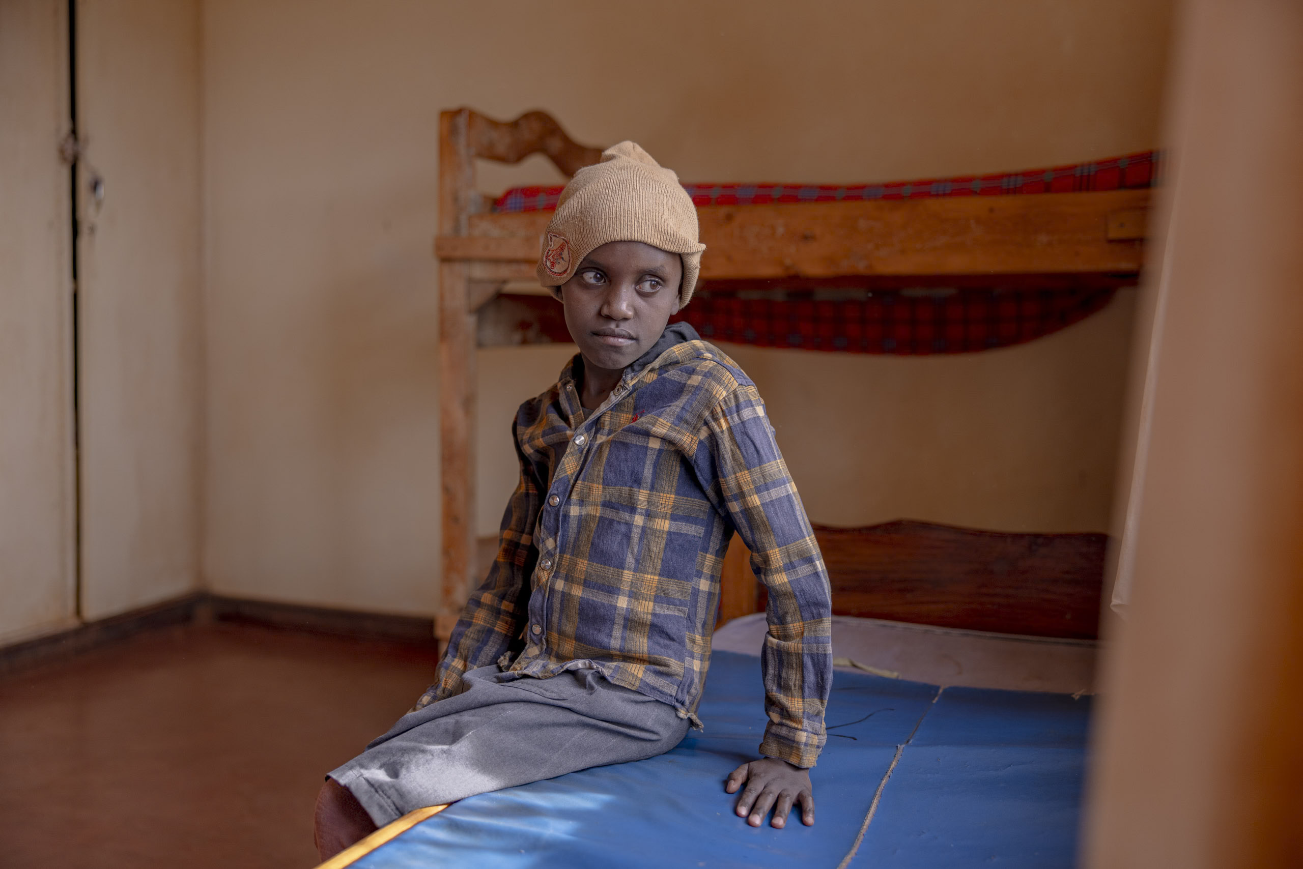Ein kleiner Junge sitzt in einem Zimmer mit einem Etagenbett aus Holz auf einer blauen Matte. Er trägt ein kariertes Hemd und eine Beanie-Mütze und blickt mit ernster Miene direkt in die Kamera. © Fotografie Tomas Rodriguez