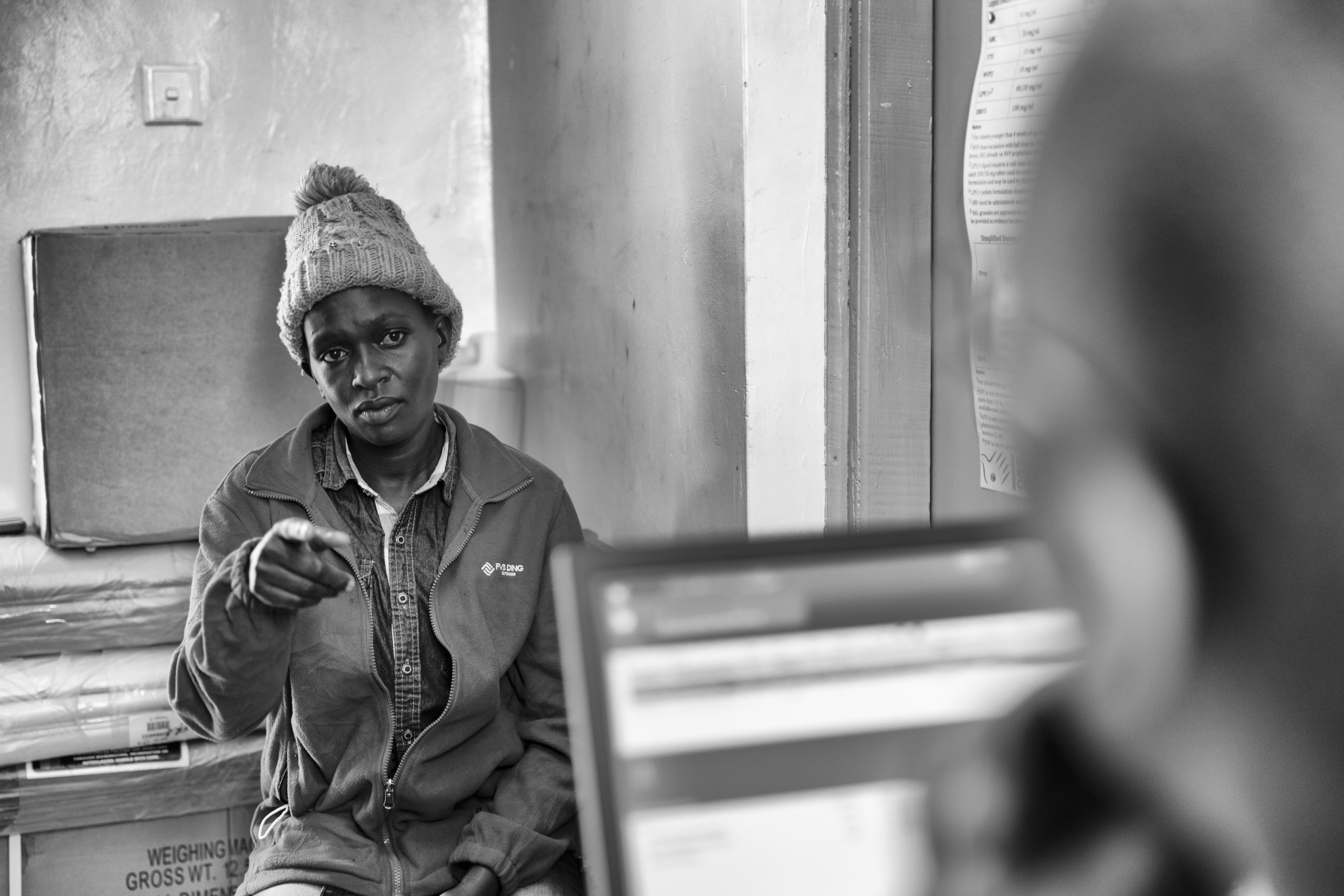 Ein Schwarzweißfoto einer Frau mit Strickmütze und Jacke, die gestikuliert, während sie mit einer Person spricht, deren Schulter und Laptop im Vordergrund zu sehen sind. Sie befinden sich in einem Innenbereich, wahrscheinlich in einem Büro. © Fotografie Tomas Rodriguez