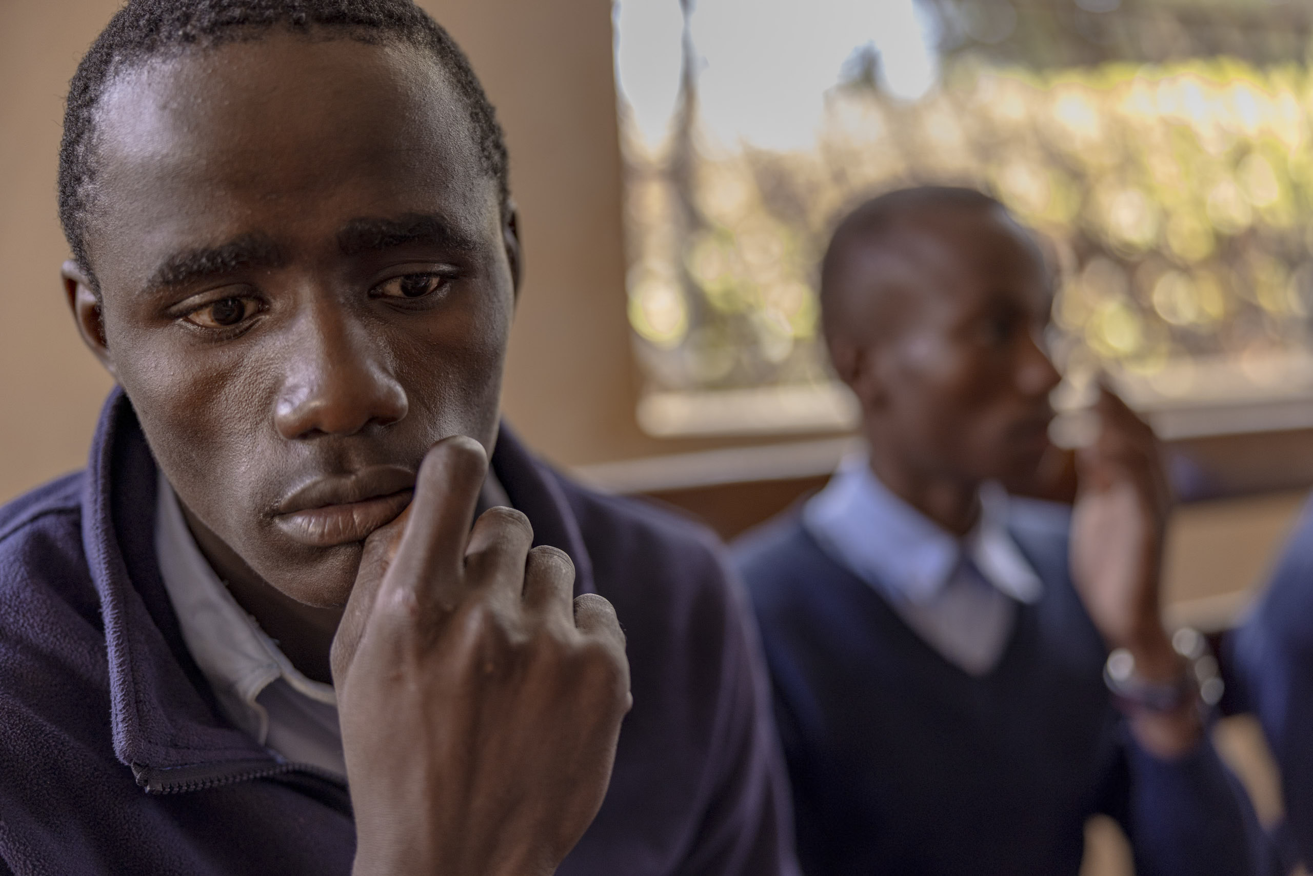 Ein nachdenklicher junger Mann mit kontemplativem Gesichtsausdruck stützt sein Kinn auf seinen Finger in einem Klassenzimmer, im Hintergrund ist eine weitere Person unscharf zu sehen. © Fotografie Tomas Rodriguez