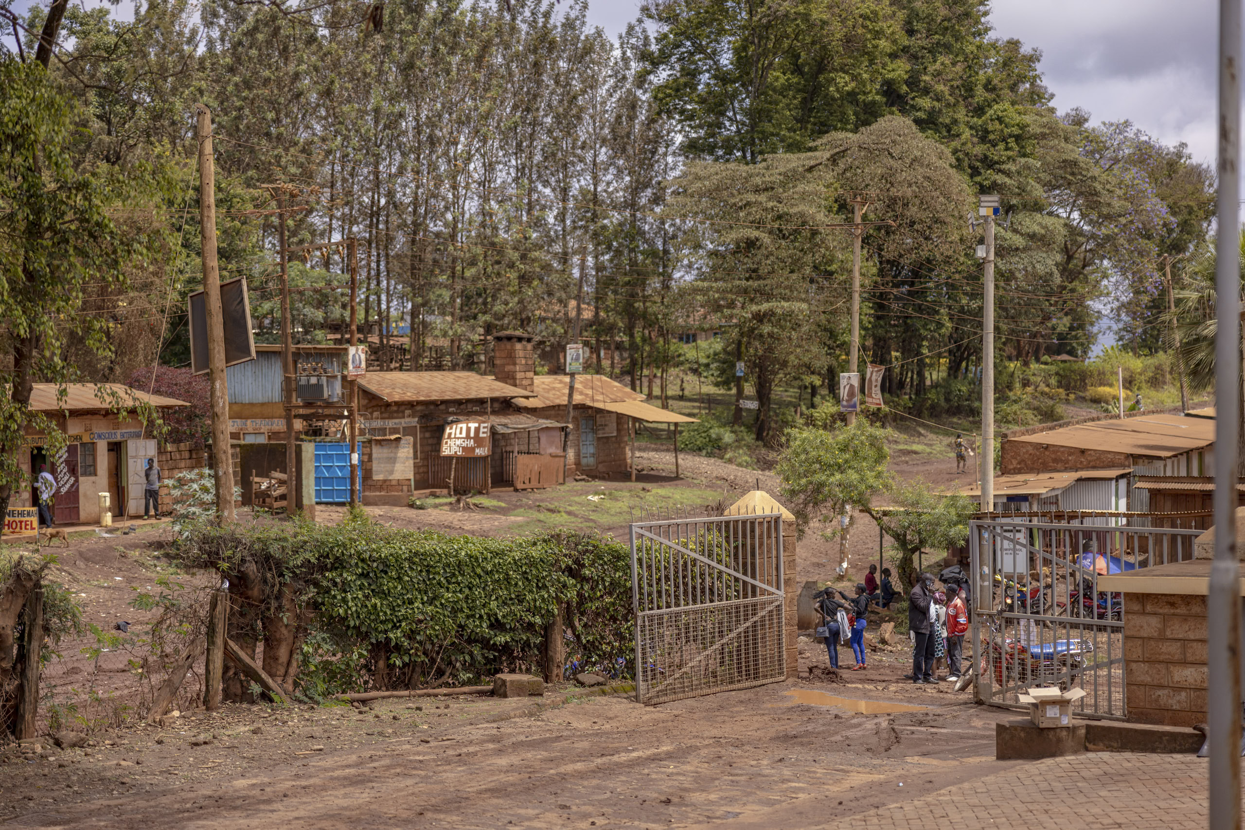 Eine rustikale Straßenszene in einer ländlichen Gegend mit einer Gruppe von Leuten, die sich in der Nähe eines Metalltors unterhalten, umgeben von kleinen Holzgebäuden und üppigen grünen Bäumen. © Fotografie Tomas Rodriguez