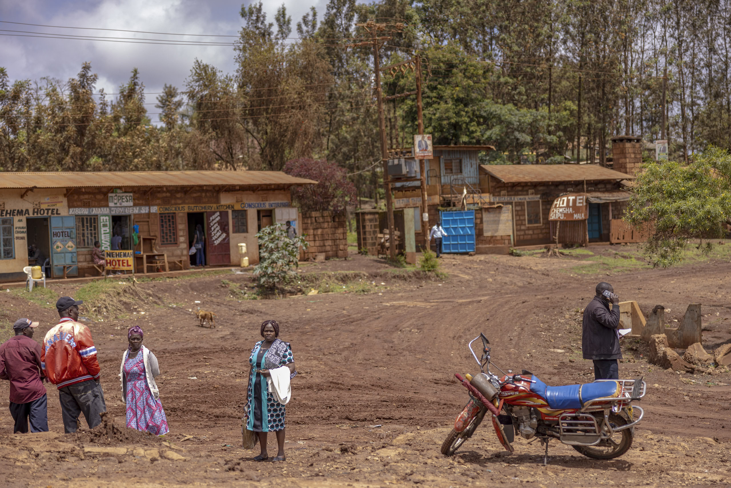 Eine ländliche Straßenszene in Kenia. Mehrere Menschen stehen und unterhalten sich in der Nähe von Geschäften. Im Vordergrund ist ein Motorrad zu sehen, im Hintergrund Bäume. © Fotografie Tomas Rodriguez