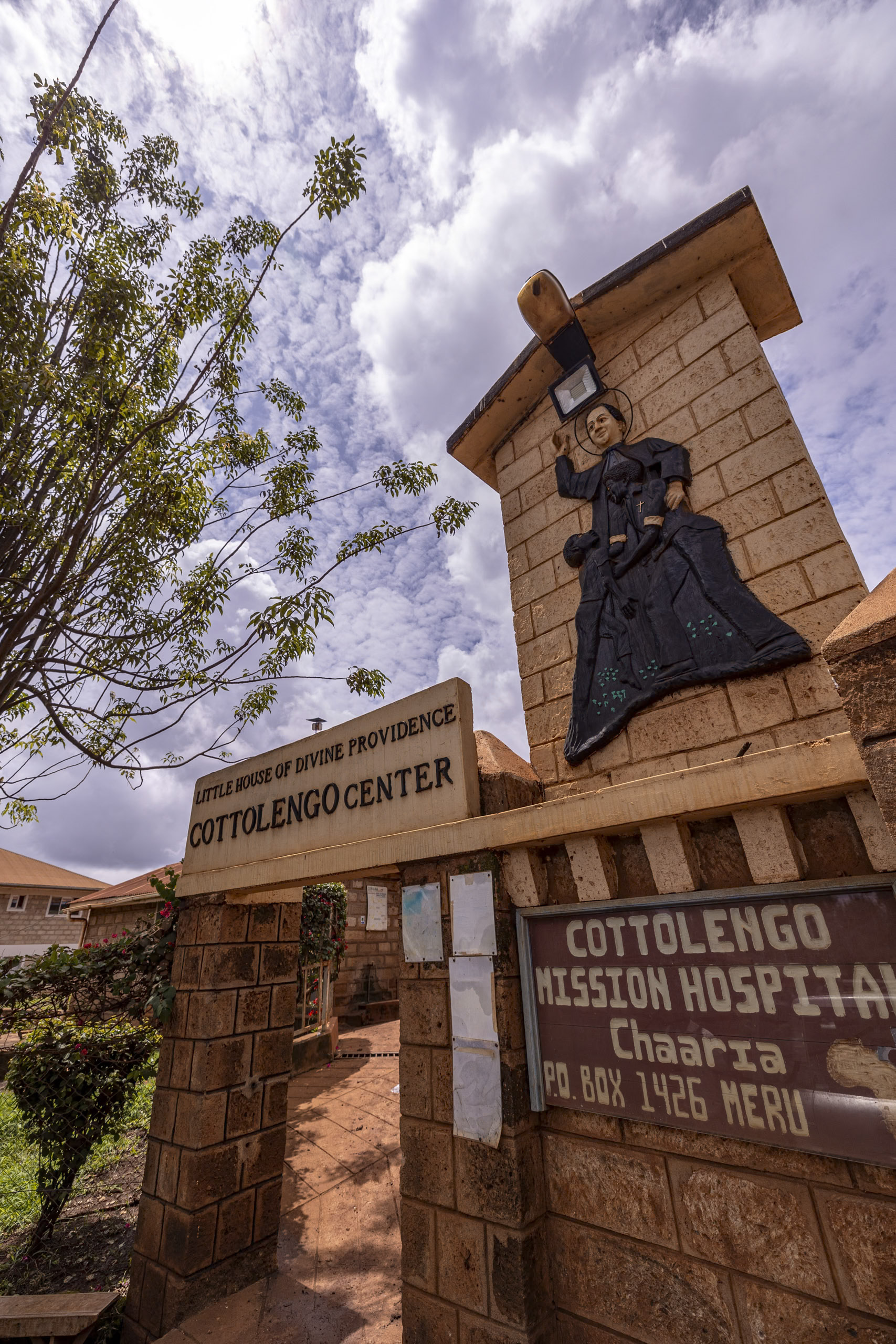 Eingang des Cotolengo-Zentrums mit der Statue einer Nonne an der Seite einer Steinsäule, unter einem blauen Himmel mit Wolken. Die Beschilderung weist darauf hin, dass es sich um ein Missionskrankenhaus handelt. © Fotografie Tomas Rodriguez