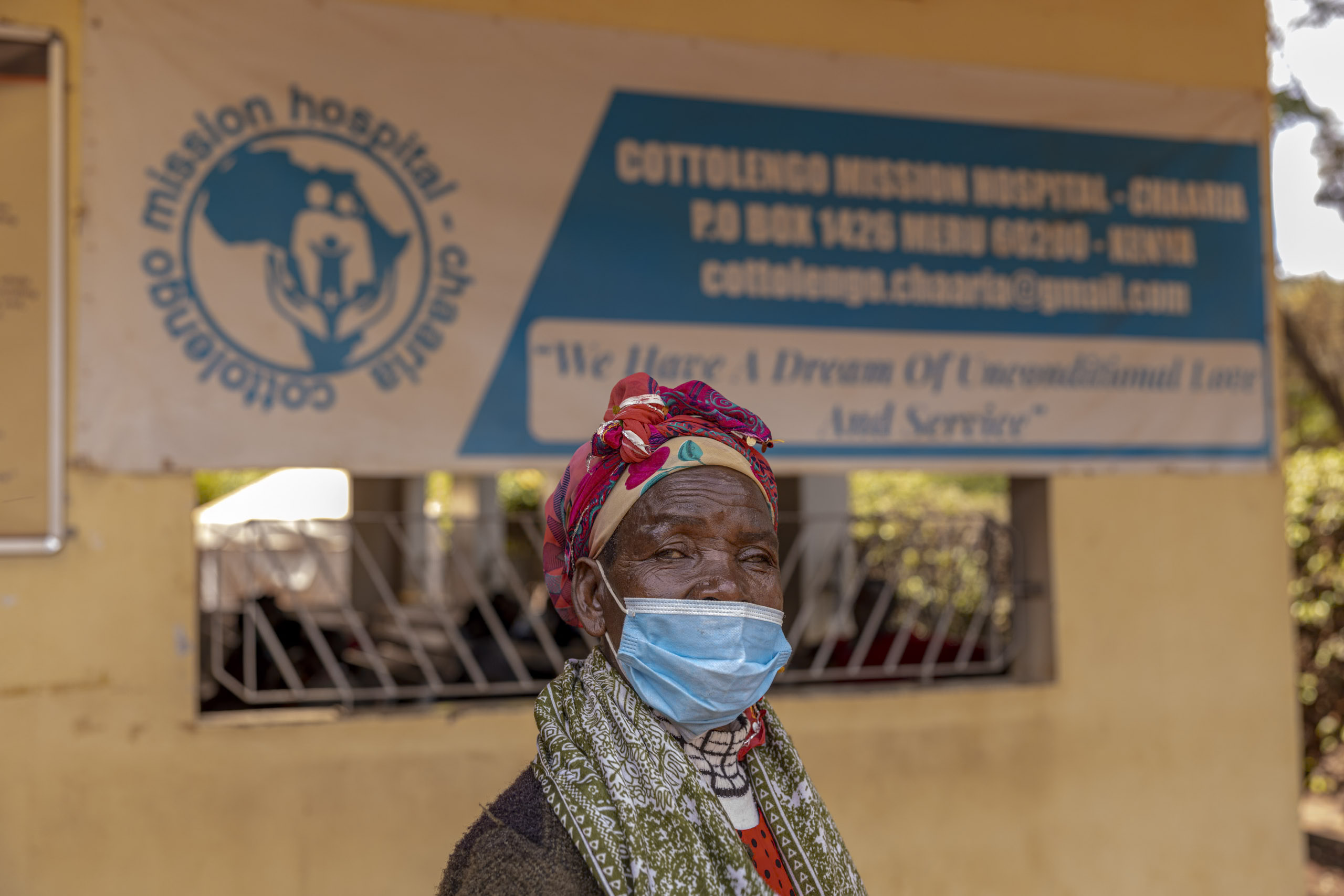 Eine ältere Frau mit buntem Kopftuch und Gesichtsmaske steht vor dem Cottolengo-Missionskrankenhaus, über ihr ist ein Schild mit Informationen zum Krankenhaus angebracht. © Fotografie Tomas Rodriguez
