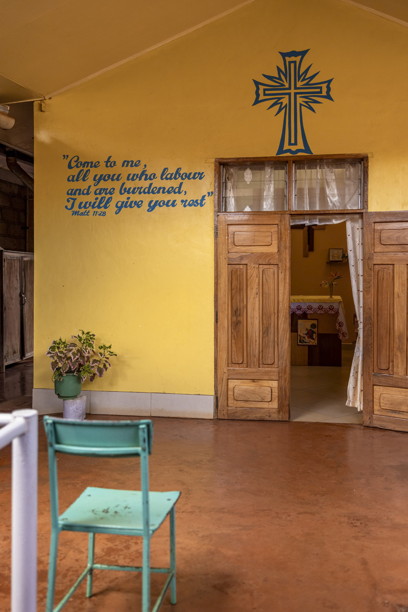 Innenansicht einer Kirche mit warmen gelben Wänden, einem leuchtend blauen Kreuz und einem Bibelzitat aus Matthäus 11:28. Holztüren führen zu einem kleinen Altar und im Vordergrund ist ein hellgrüner Stuhl zu sehen. © Fotografie Tomas Rodriguez