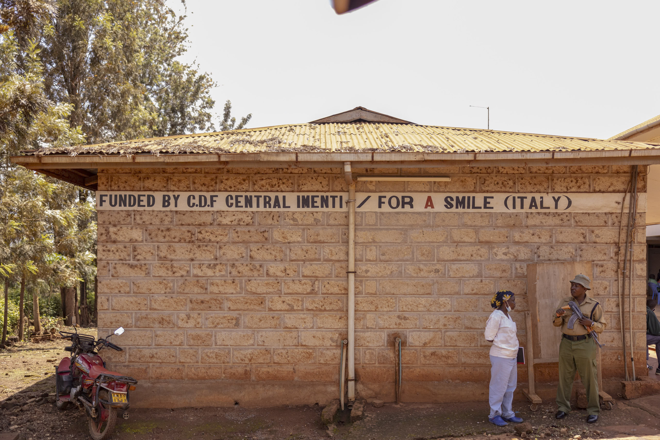 Ein kleines Gebäude aus Ziegeln mit einem Schild „finanziert von CDF Central Inetni / Für ein Lächeln (Italien)“. Zwei Personen unterhalten sich draußen, während eine dritte mit einem Motorrad in der Nähe steht. © Fotografie Tomas Rodriguez
