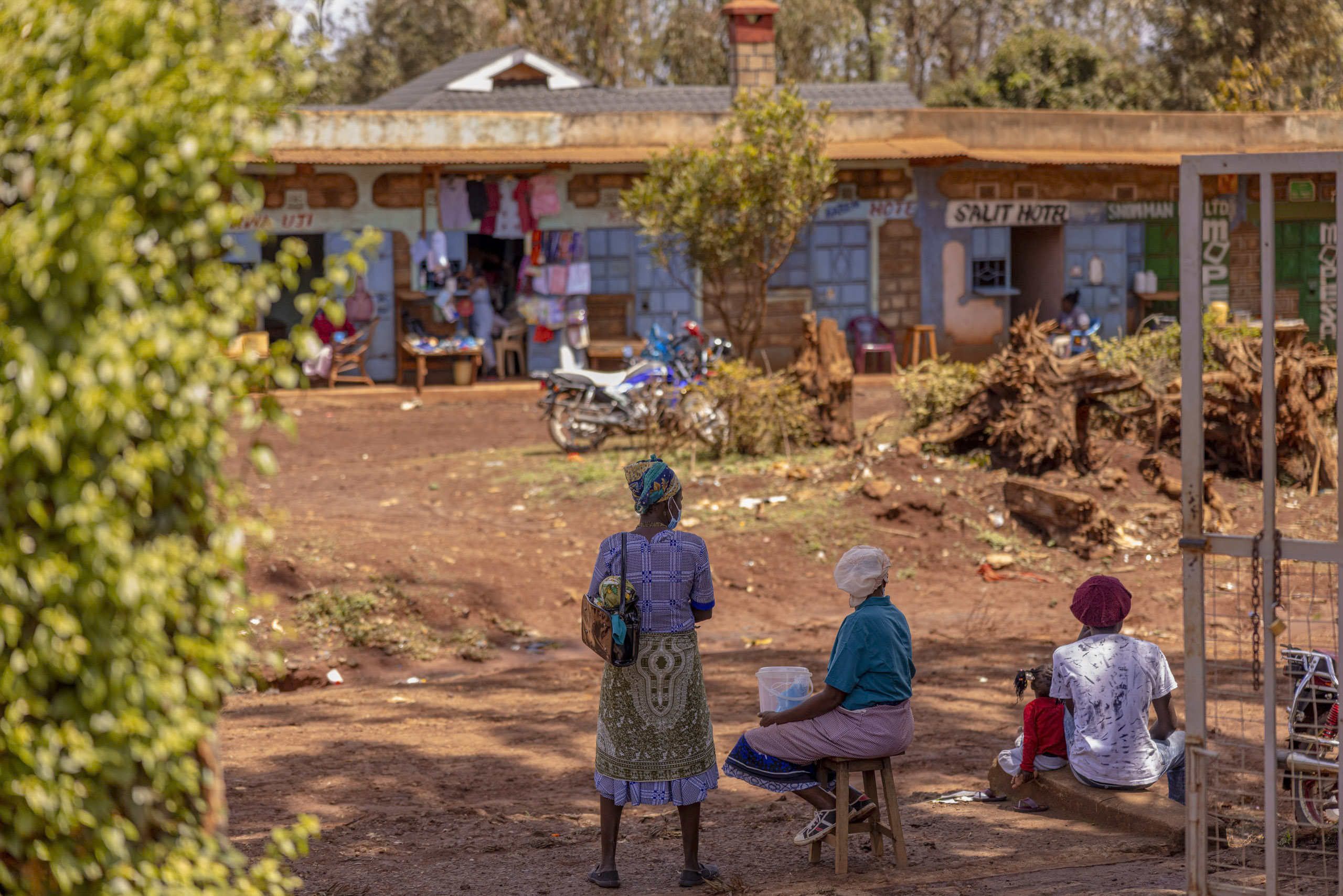 Ländliche afrikanische Szene mit Menschen, die in der Nähe eines farbenfrohen Marktes mit Schildern, einem geparkten Motorrad und vor den Geschäften hängender Kleidung sitzen und stehen. Eine ruhige, alltägliche Umgebung. © Fotografie Tomas Rodriguez