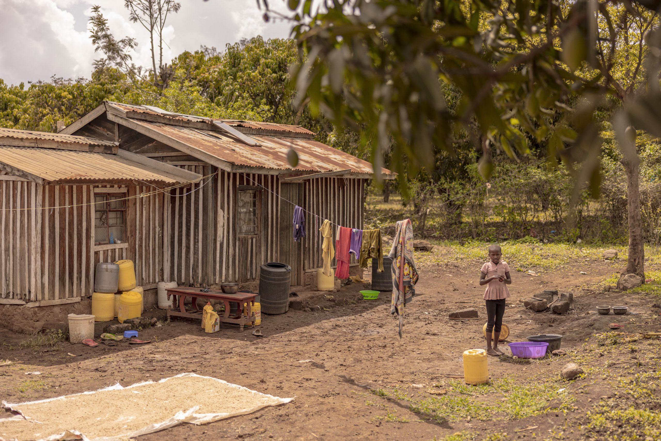 Ein Kind steht vor einem rustikalen Haus mit Wellblechwänden und Blechdach, umgeben von trocknendem Getreide, hängender Wäsche und verschiedenen Haushaltsgegenständen in einer ländlichen Landschaft. © Fotografie Tomas Rodriguez