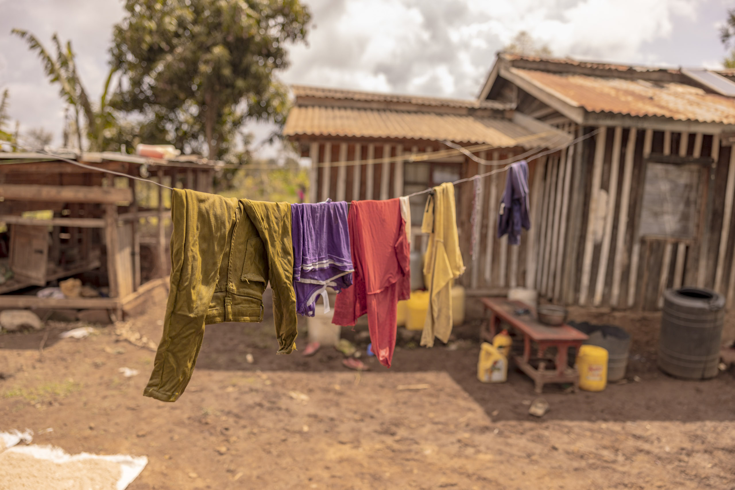 Bunte Kleidung hängt zum Trocknen auf einer Leine in einem ländlichen Hinterhof mit rustikalen Gebäuden und einem bewölkten Himmel im Hintergrund. © Fotografie Tomas Rodriguez