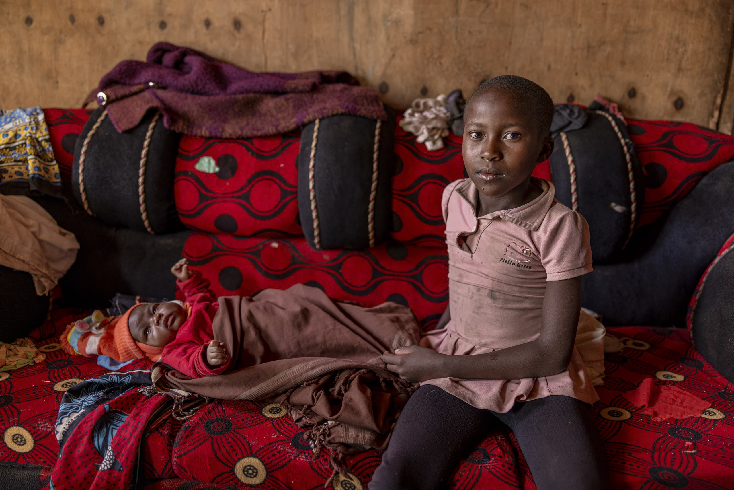 Ein junges Mädchen sitzt neben einem schlafenden Baby in einem schwach beleuchteten Raum, der durch bunte Kissen und Decken hervorgehoben wird. Das Mädchen blickt mit ernster Miene direkt in die Kamera. © Fotografie Tomas Rodriguez