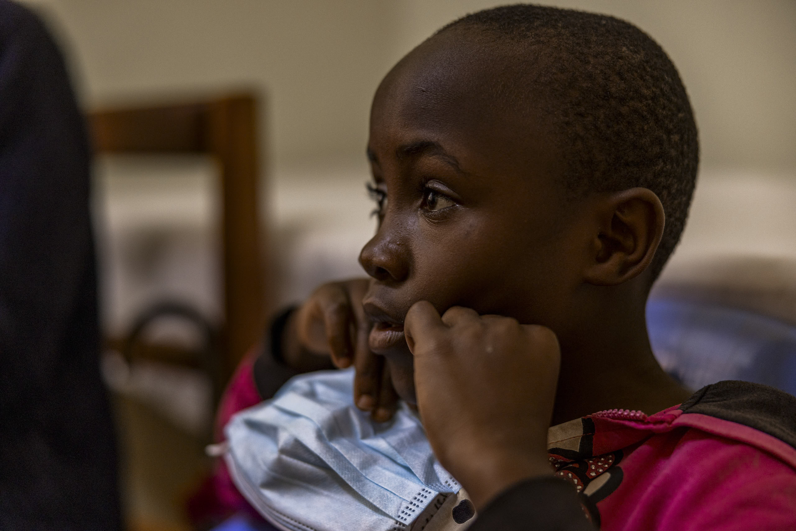 Eine Nahaufnahme eines jungen afrikanischen Kindes, das in einem schwach beleuchteten Innenraum aufmerksam zur Seite blickt, die Hände vor dem Gesicht hält und ein rosa und schwarz gestreiftes Hemd trägt. © Fotografie Tomas Rodriguez
