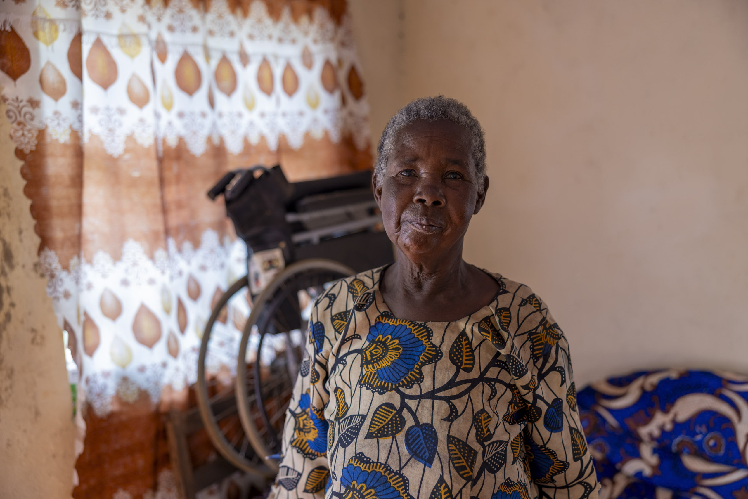 Eine ältere Frau mit sanftem Gesichtsausdruck steht in einem Raum mit traditioneller Einrichtung, einschließlich gemusterten Vorhängen. Im Hintergrund ist ein Rollstuhl zu sehen. © Fotografie Tomas Rodriguez