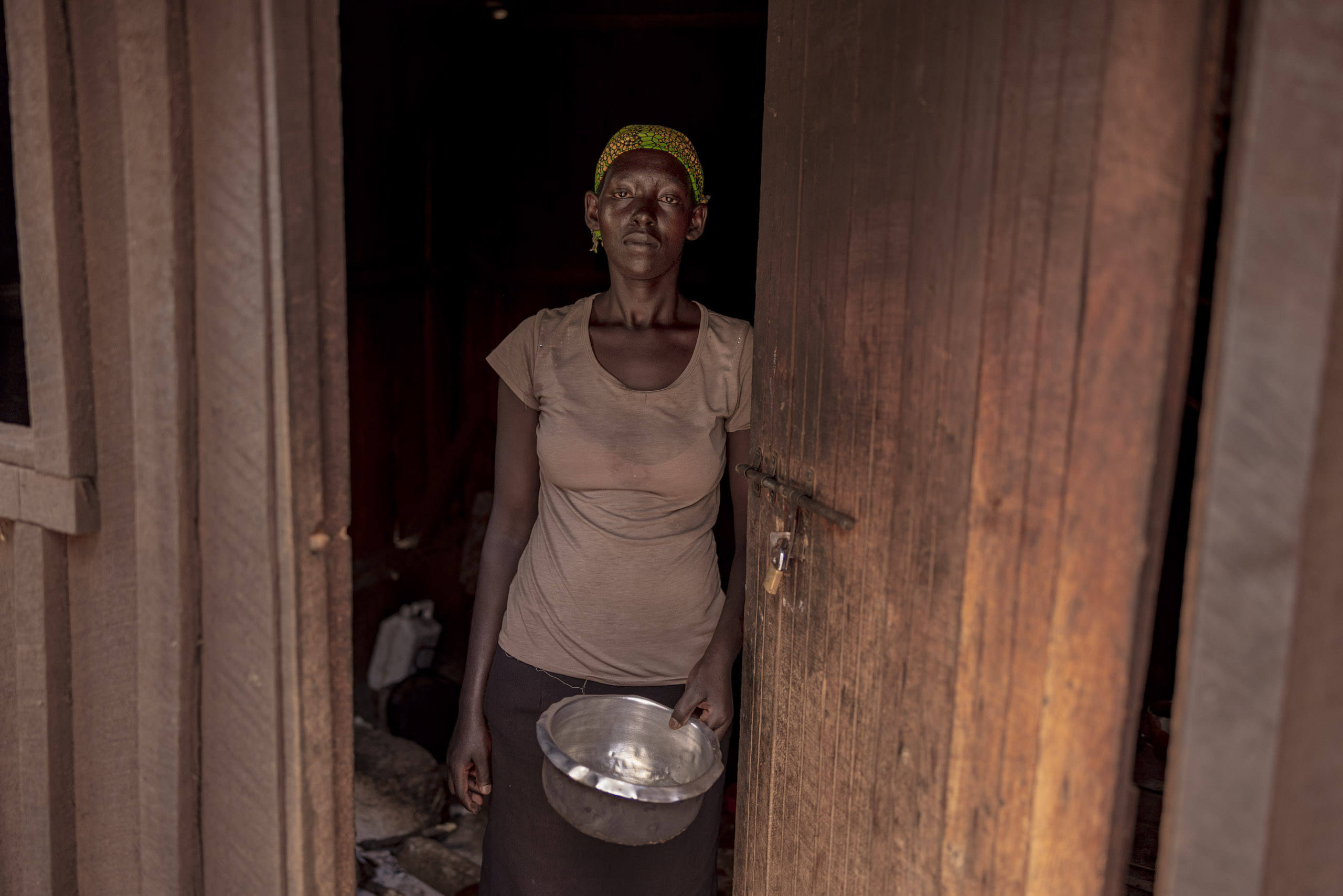 Eine Frau steht mit ernster Miene in einer Tür, hält eine Metallschüssel in der Hand. Sie trägt ein braunes Oberteil und ein buntes Stirnband. Der Hintergrund ist schwach beleuchtet, was ihre Präsenz im Bild betont. © Fotografie Tomas Rodriguez