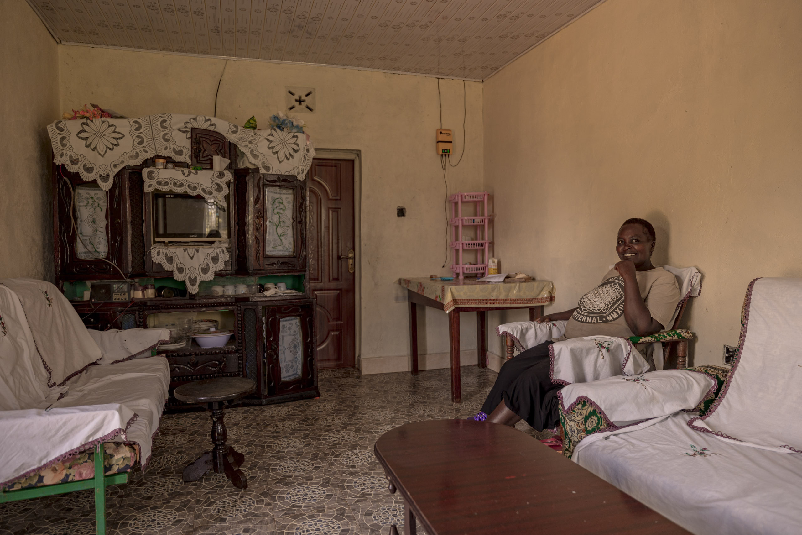 Eine Frau lächelt, während sie auf einem Sofa in einem traditionellen Wohnzimmer mit kunstvollen Holzmöbeln, dekorativen Spiegeln und gefliestem Boden sitzt. © Fotografie Tomas Rodriguez