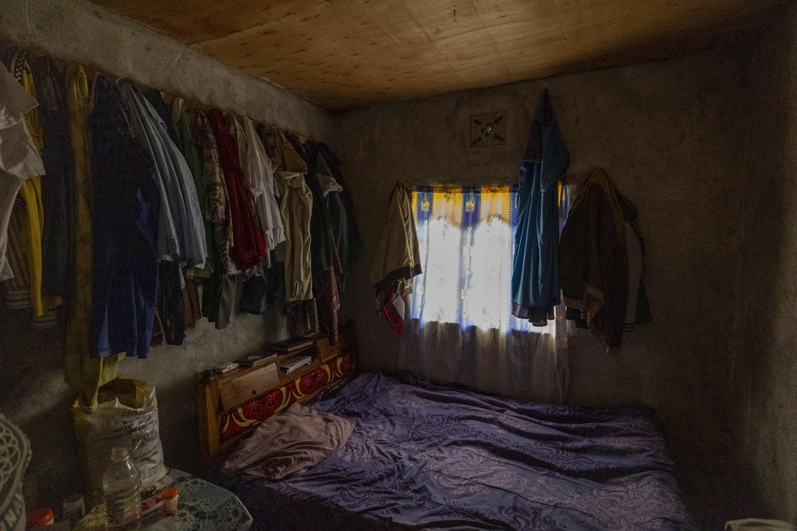Ein schwach beleuchteter Raum, in dem links Kleidung hängt, rechts ein Bett mit einer lila Decke steht und unter einem Fenster mit gelben Vorhängen eine kleine Kommode steht. © Fotografie Tomas Rodriguez