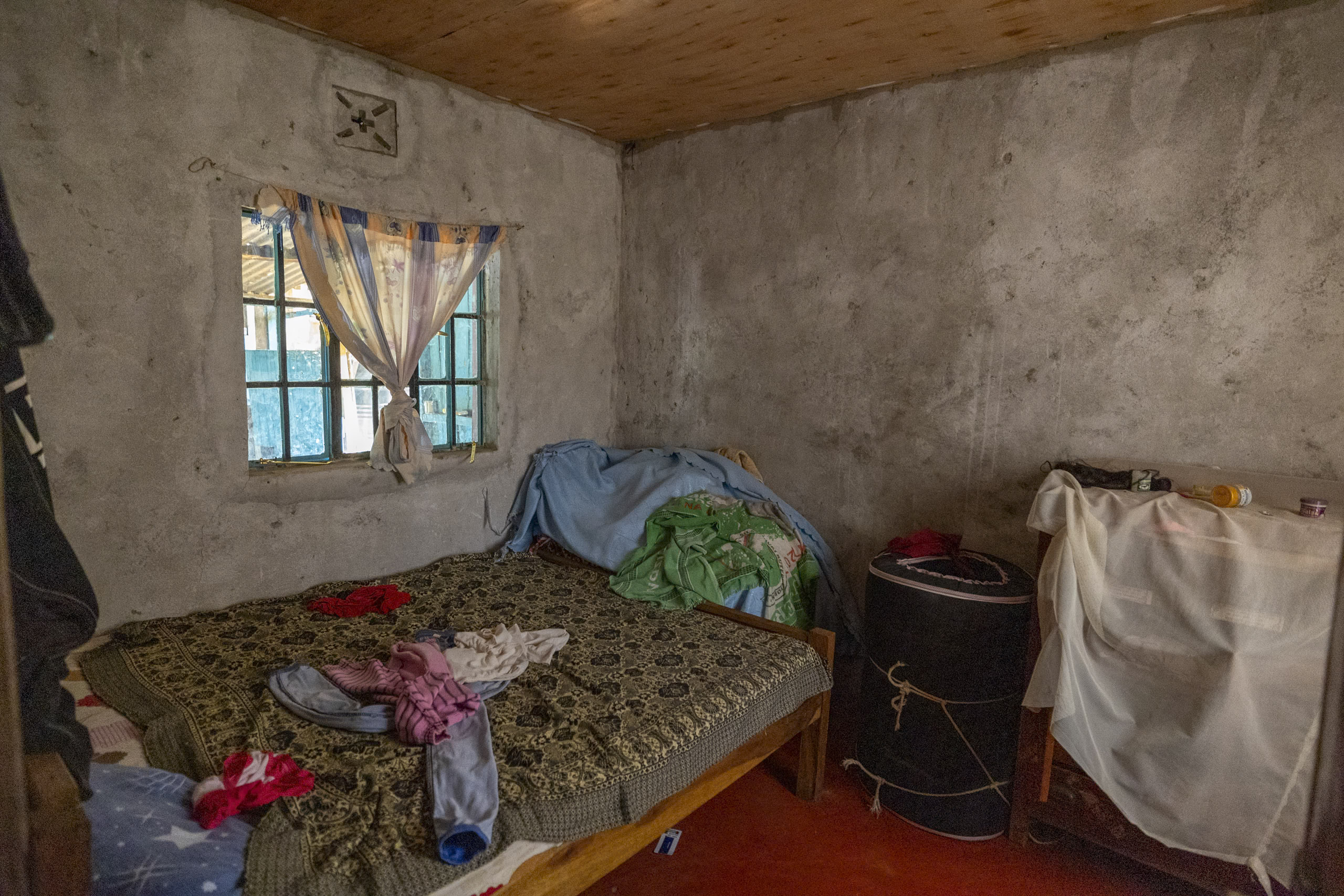 Ein bescheidenes, rustikales Schlafzimmer mit einem Einzelbett, das mit einer gemusterten Decke und verstreuten Kleidungsstücken bedeckt ist, einer Betonwand, einem kleinen Fenster mit Vorhängen und einfacher Einrichtung. © Fotografie Tomas Rodriguez