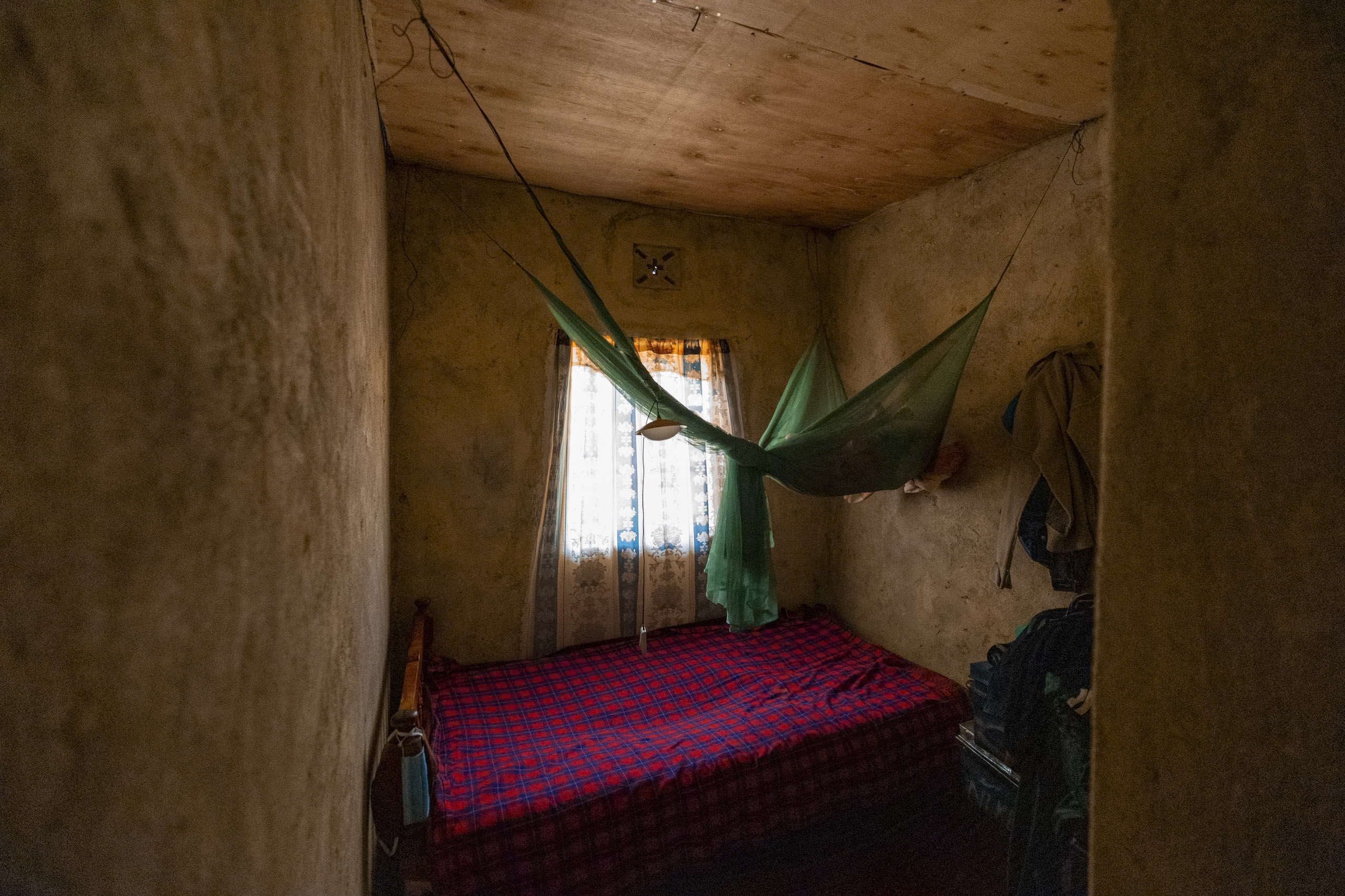 Ein kleines, rustikales Zimmer mit einer grünen Hängematte über einem rot und blau gemusterten Bett. Ein einzelnes Fenster lässt Licht herein, erhellt die nackten Holzwände und schafft eine schlichte, schwach beleuchtete Atmosphäre. © Fotografie Tomas Rodriguez