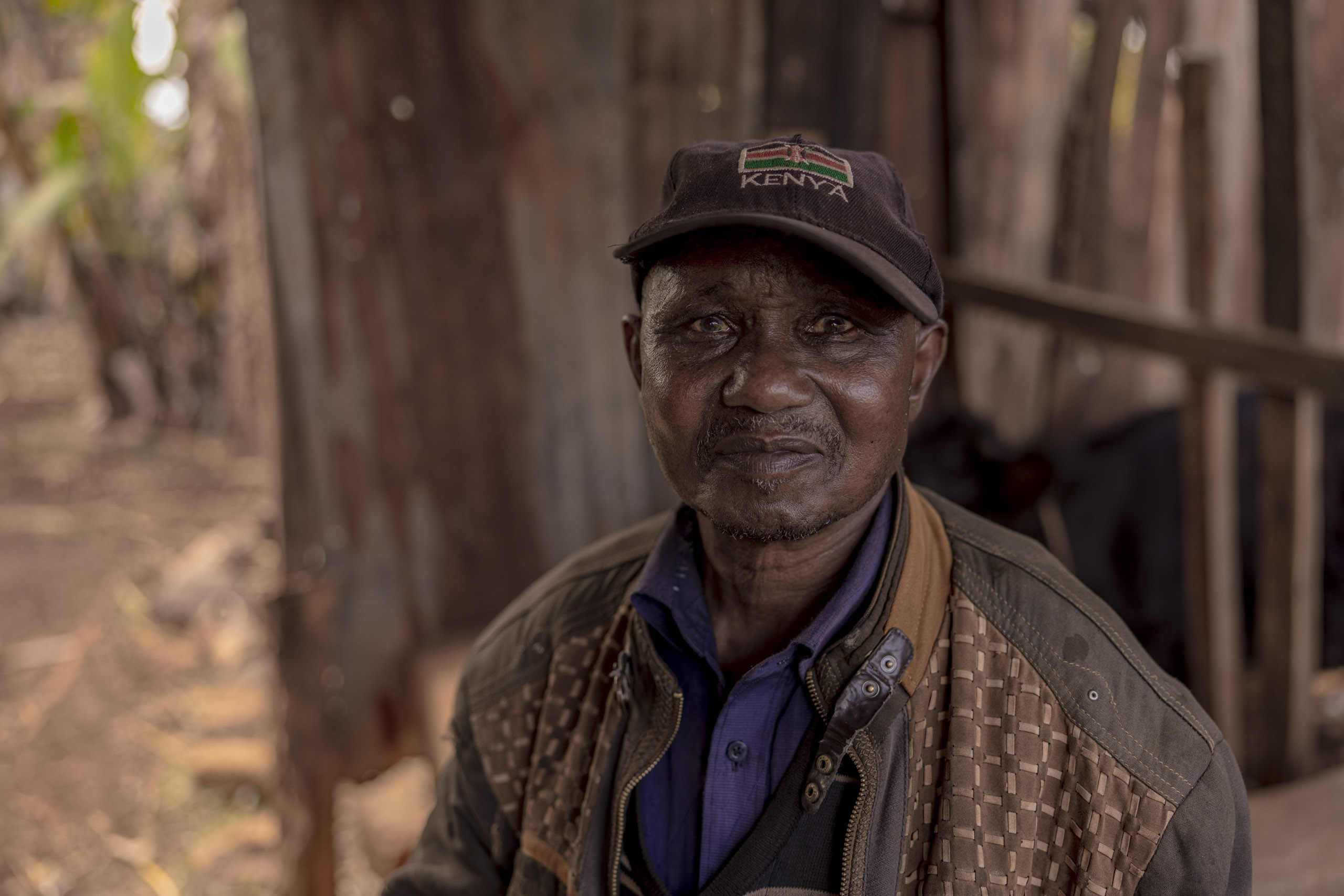 Ein älterer Mann mit „Kenia“-Mütze und Steppjacke lächelt sanft und sitzt vor einer rustikalen Holzkulisse. © Fotografie Tomas Rodriguez