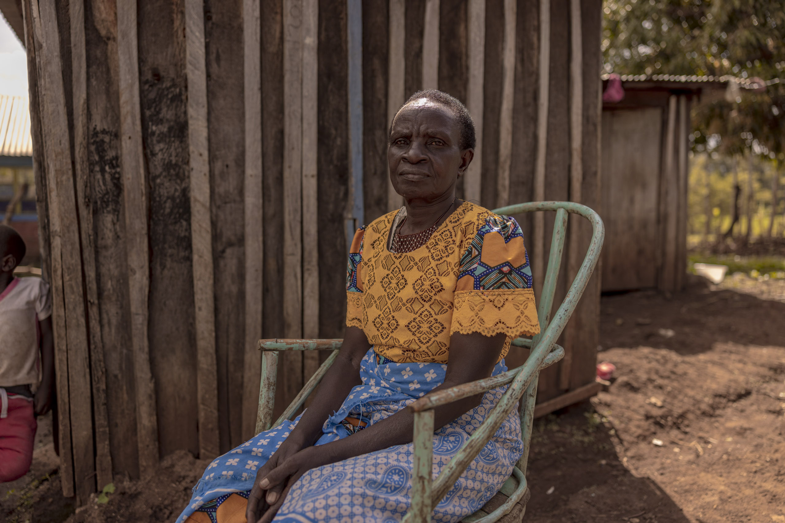 Eine ältere Afrikanerin in farbenfroher Tracht sitzt im Freien auf einem Metallstuhl, im Hintergrund sind Holzlatten und ein kleines Kind zu sehen. © Fotografie Tomas Rodriguez