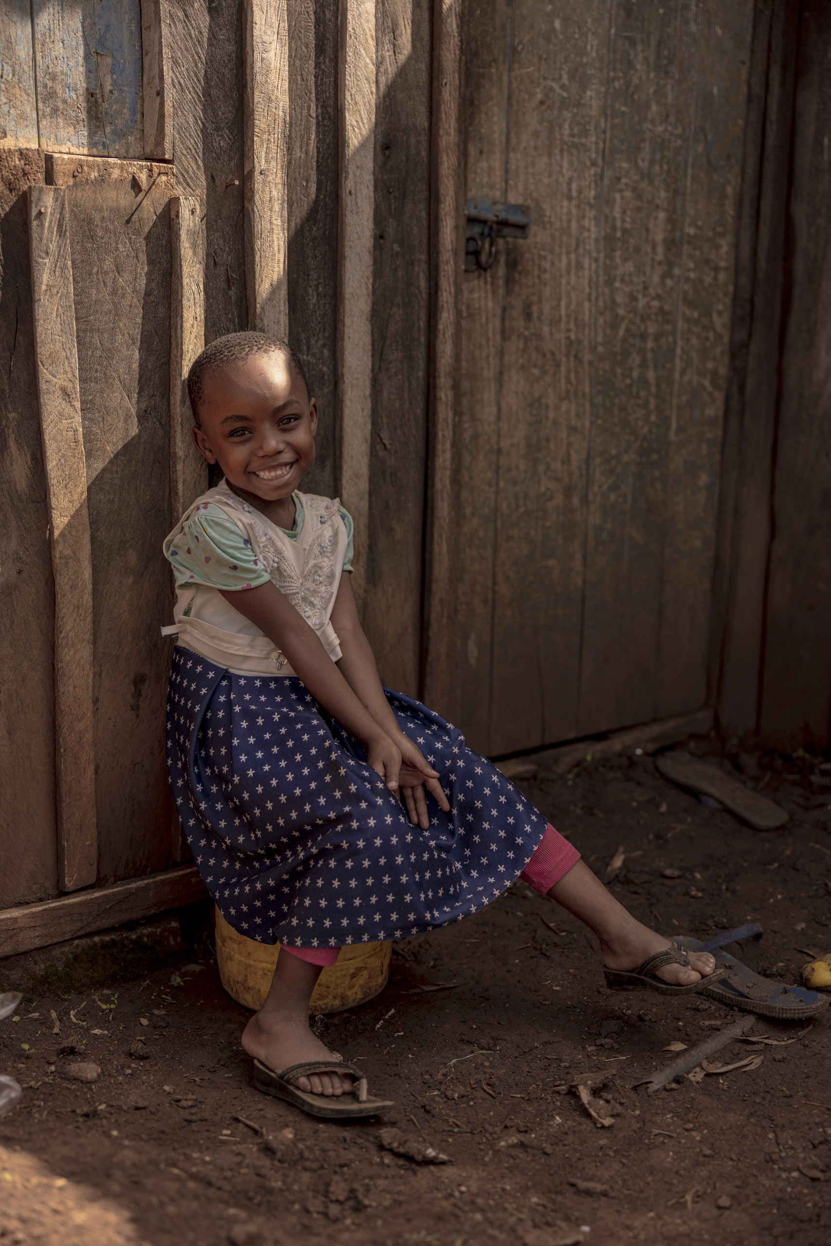 Ein junges Mädchen mit einem fröhlichen Lächeln hockt in einer sonnigen, rustikalen Umgebung vor einer Holztür, bekleidet mit einem gepunkteten Rock und Flip-Flops. © Fotografie Tomas Rodriguez