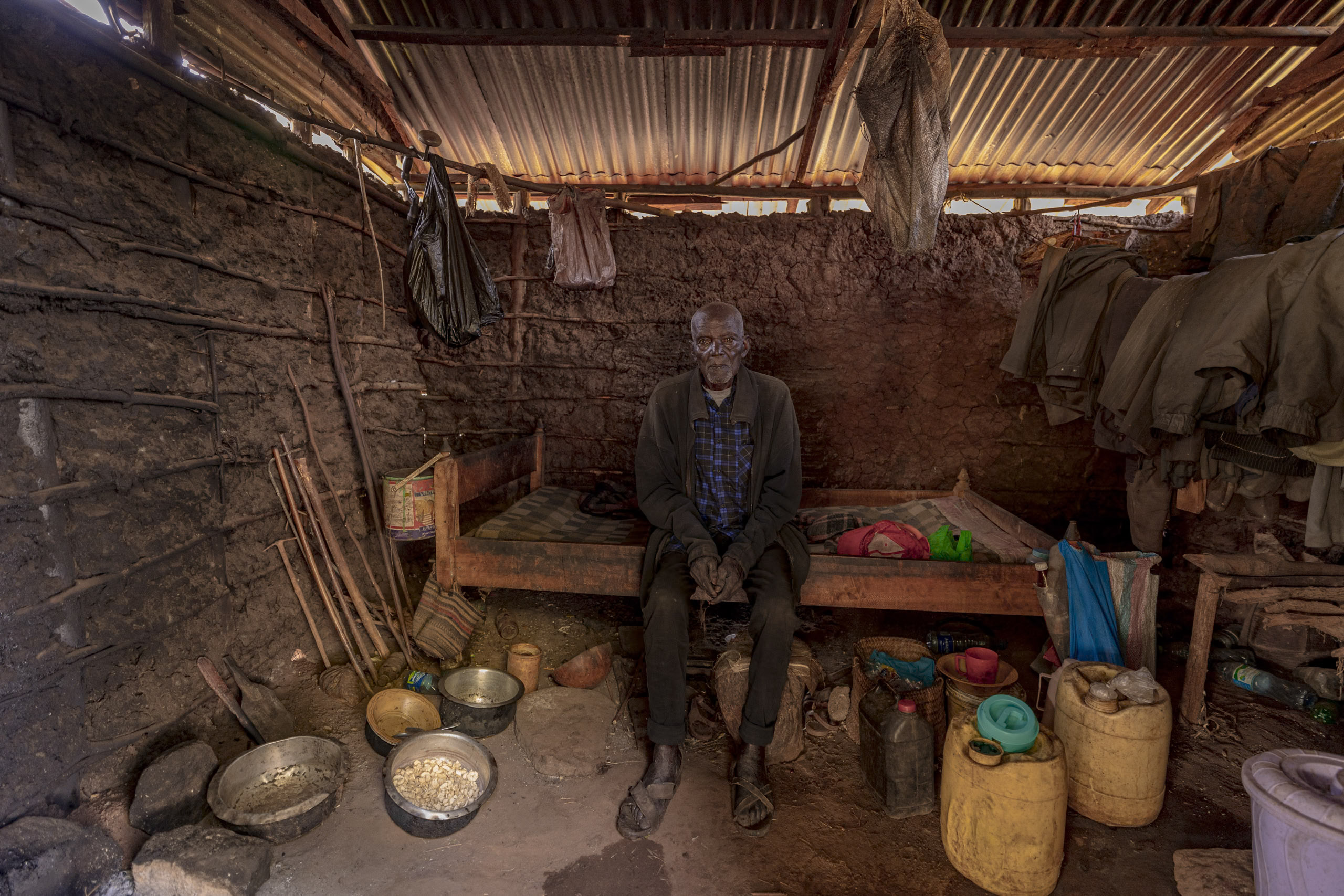 Ein älterer Mann sitzt auf einer Bank in einer rustikalen Küche mit Lehmwänden und einem Wellblechdach. Verschiedene Töpfe, Utensilien und Behälter sind im Raum verstreut. © Fotografie Tomas Rodriguez