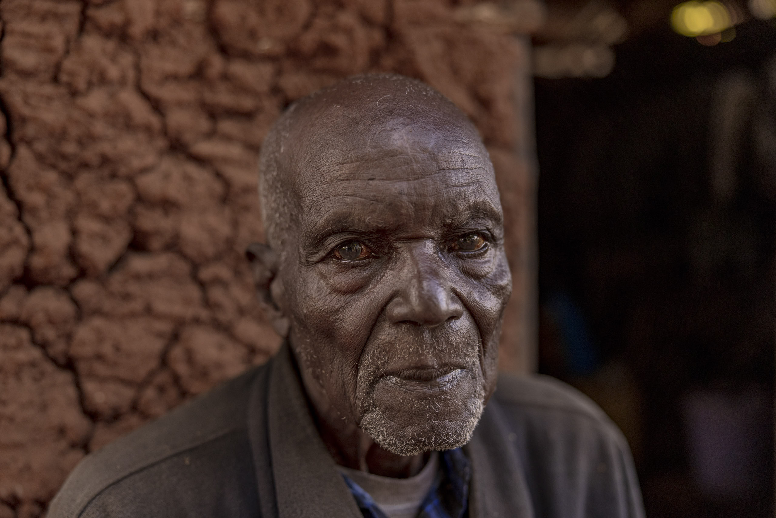 Ein älterer Mann mit tief zerfurchtem Gesicht und nachdenklichen Augen blickt direkt in die Kamera und posiert vor dem unscharfen Hintergrund einer Lehmmauer. Er trägt eine graue Jacke über einem blauen Hemd. © Fotografie Tomas Rodriguez