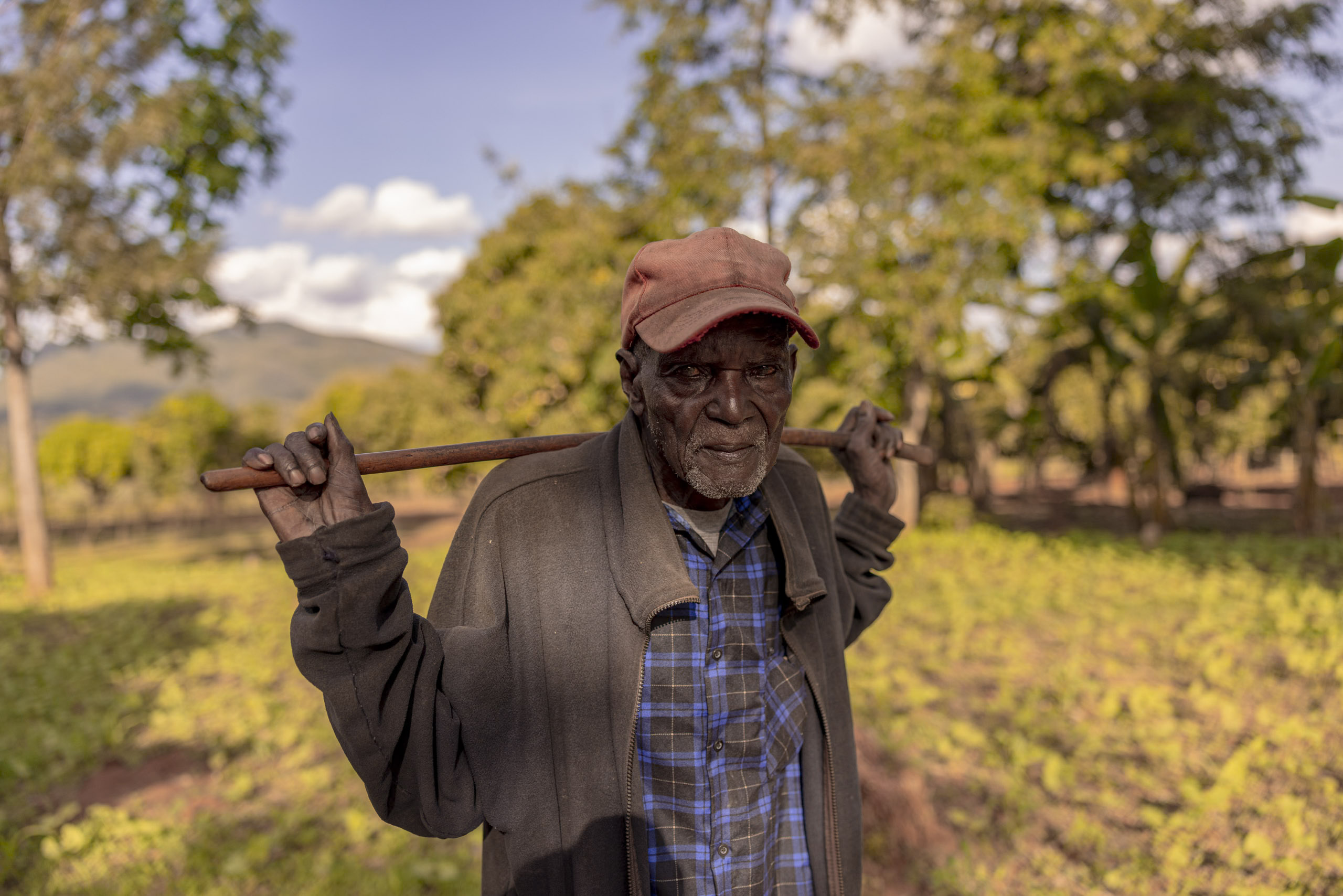 Ein älterer Mann mit wettergegerbtem Gesicht, einer roten Mütze und einer dunklen Jacke trägt eine Hacke über der Schulter in einer ländlichen Landschaft mit Bäumen und Hügeln im Hintergrund. © Fotografie Tomas Rodriguez