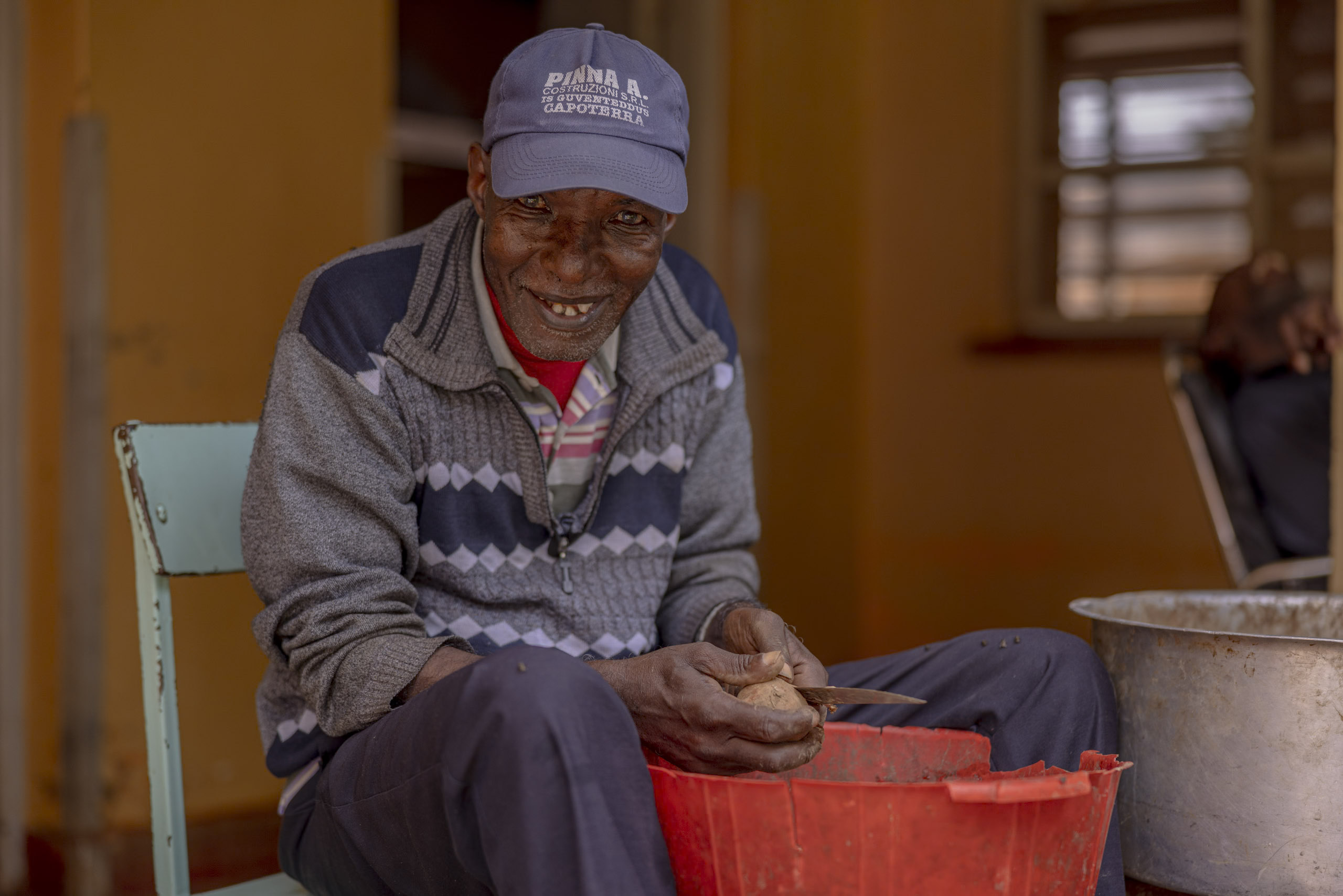 Ein älterer Mann in Pullover und Mütze sitzt auf einem Stuhl und schält Kartoffeln in einen roten Eimer, während er in einer rustikalen Umgebung herzlich in die Kamera lächelt. © Fotografie Tomas Rodriguez